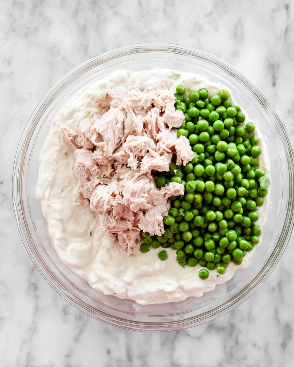 A clear glass bowl sits on a white marbled surface. Inside the bowl, there is one base layer of thick white creamy sauce spread evenly. On top of this base, there are two separate piles: a light pink flaky meat on the upper half and bright green frozen peas on the lower half. The textures contrast with the smooth sauce, the flaky meat chunks, and the round peas. The photo taken with an iphone --ar 4:5 --v 7
