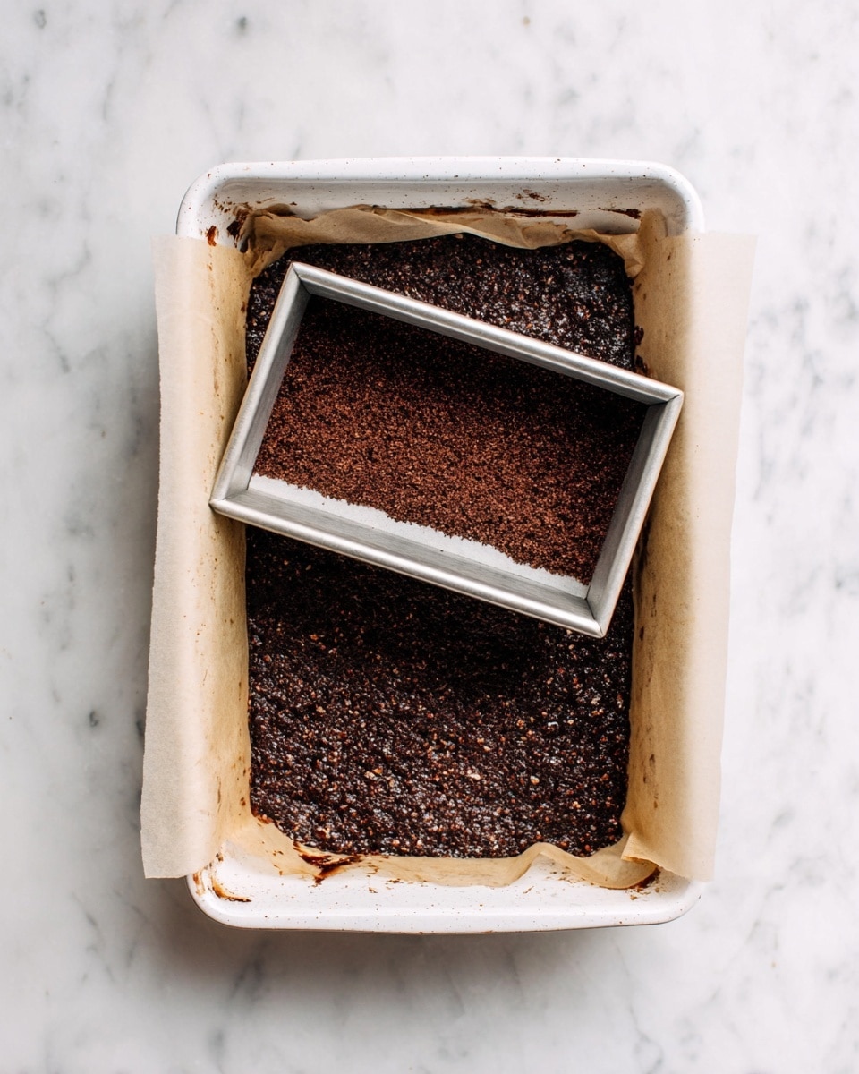 The image shows a white baking pan lined with parchment paper filled with a dark brown mixture with a rough texture, possibly a nut or chocolate-based recipe. Inside the bigger pan, there is a smaller rectangular baking pan placed upside down in the center, pressing down on the mixture to create a flat layer around it. The mixture around the smaller pan looks uneven but packed tightly, while the smaller pan has a smooth, shiny surface with light stains. The whole setup is on a white marbled surface. photo taken with an iphone --ar 4:5 --v 7
