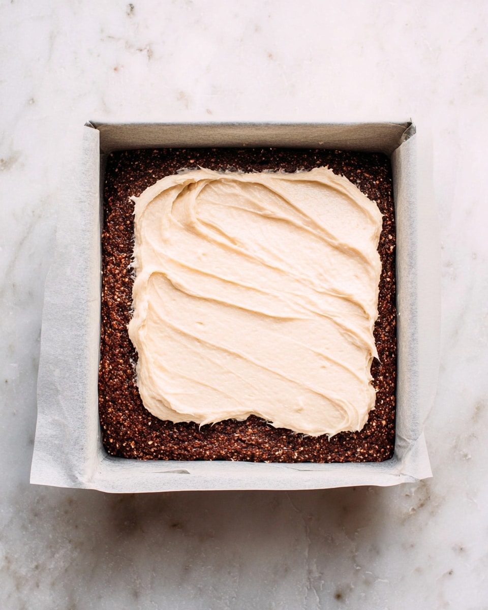 A square baking pan lined with white parchment paper holds a two-layered dessert. The bottom layer is thick, dark brown, and textured with small visible bits, suggesting a dense, baked base. On top, a creamy, light beige frosting is being spread unevenly, covering roughly half of the surface with visible swirls and a smooth, soft texture. The pan sits on a white marbled surface. photo taken with an iphone --ar 4:5 --v 7