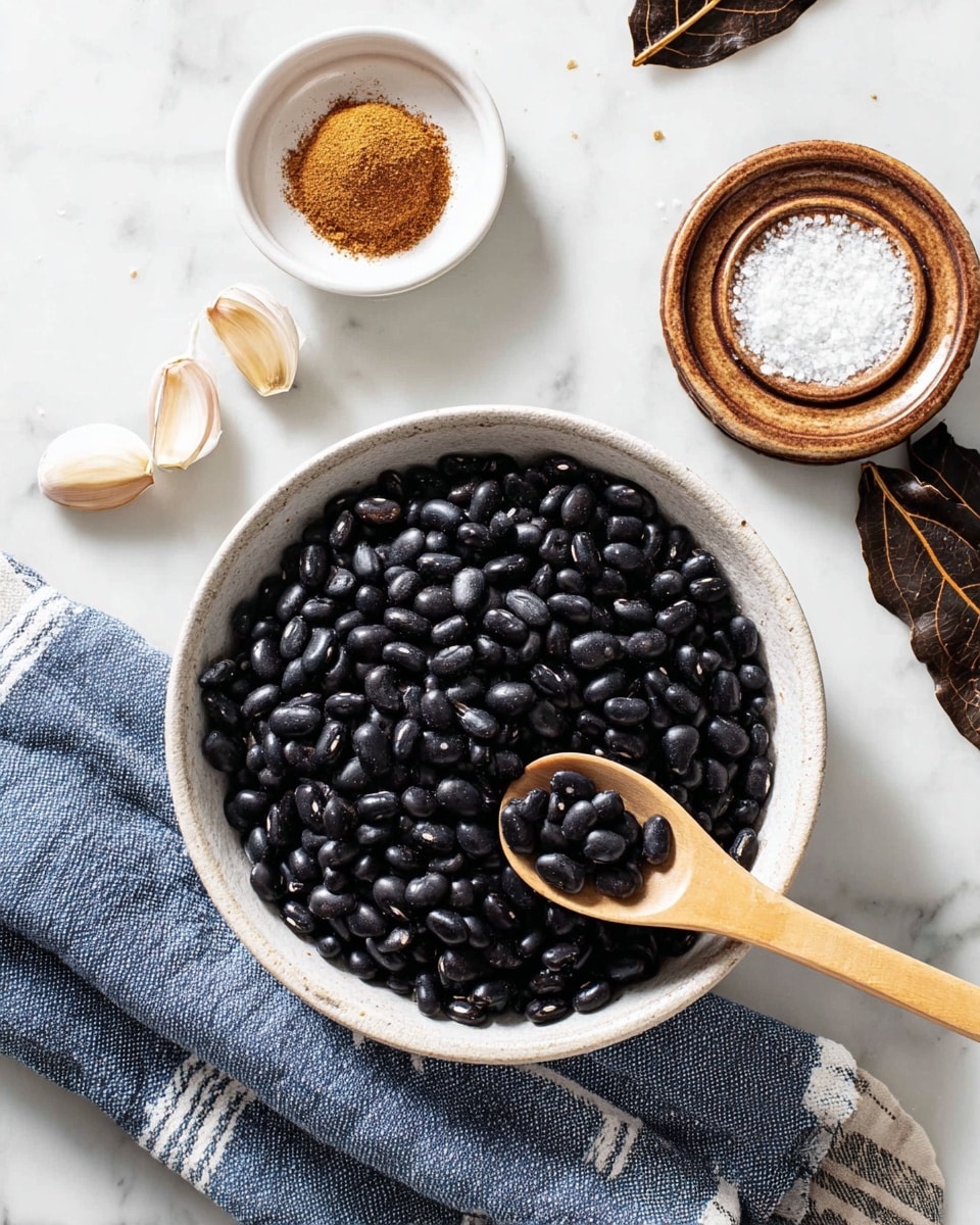 A round white bowl filled with shiny black beans sits on a white marbled surface, with a light wooden spoon resting inside the bowl, scooping some beans. Below the bowl is a blue and white striped cloth. Above the bowl, there are three garlic cloves placed on the white marbled surface, a small white dish filled with brown spice powder, and a small wooden plate with white salt arranged in a spiral shape. To the right, there are dark brown dried leaves partially visible on the white marbled surface. Photo taken with an iphone --ar 4:5 --v 7