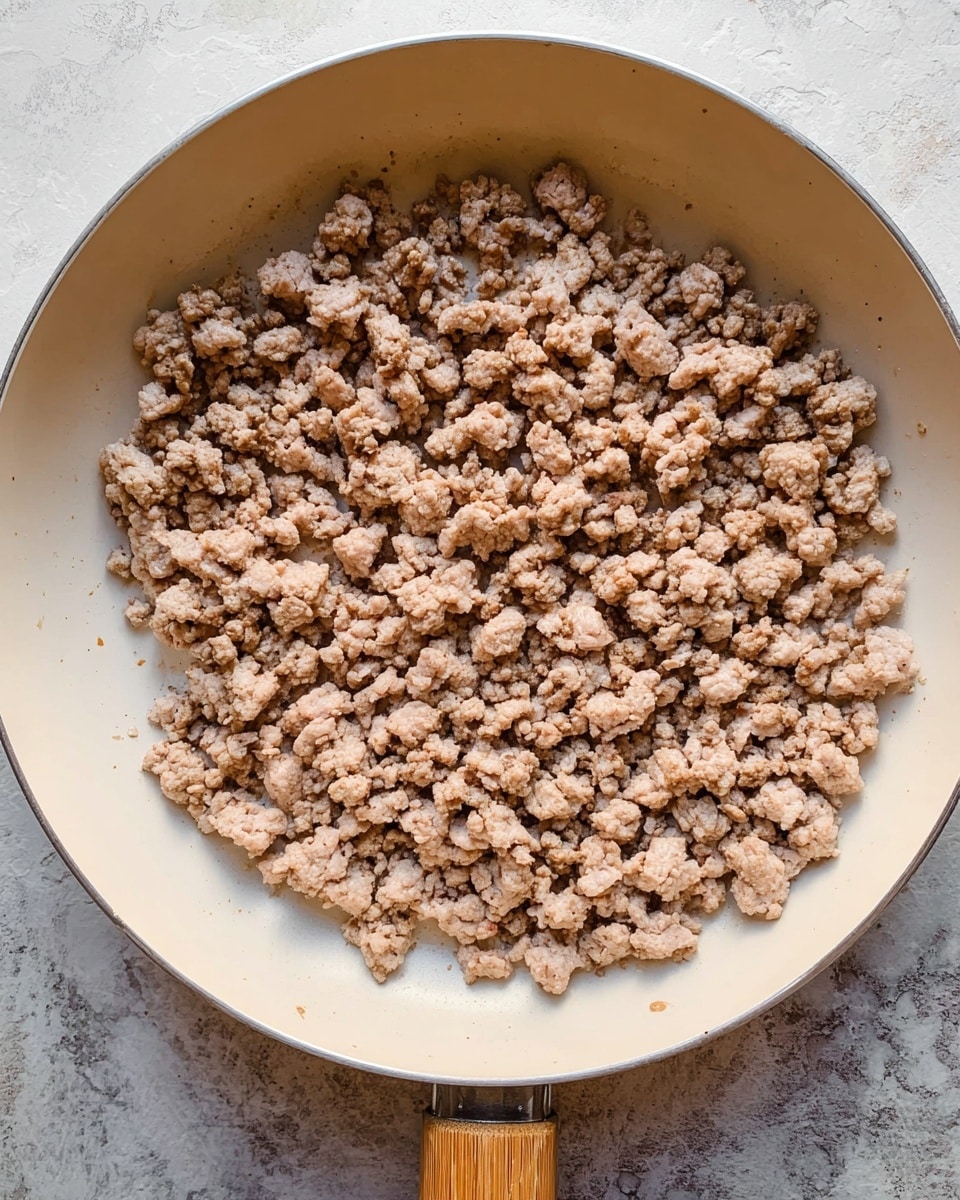 A large white frying pan filled with cooked ground meat pieces that are light brown and crumbly in texture, spread evenly across the pan's surface. The pan has a wooden handle and rests on a white marbled surface, while the meat shows slight variation in shades from pale brown to darker brown spots, indicating it is fully cooked. Photo taken with an iphone --ar 4:5 --v 7