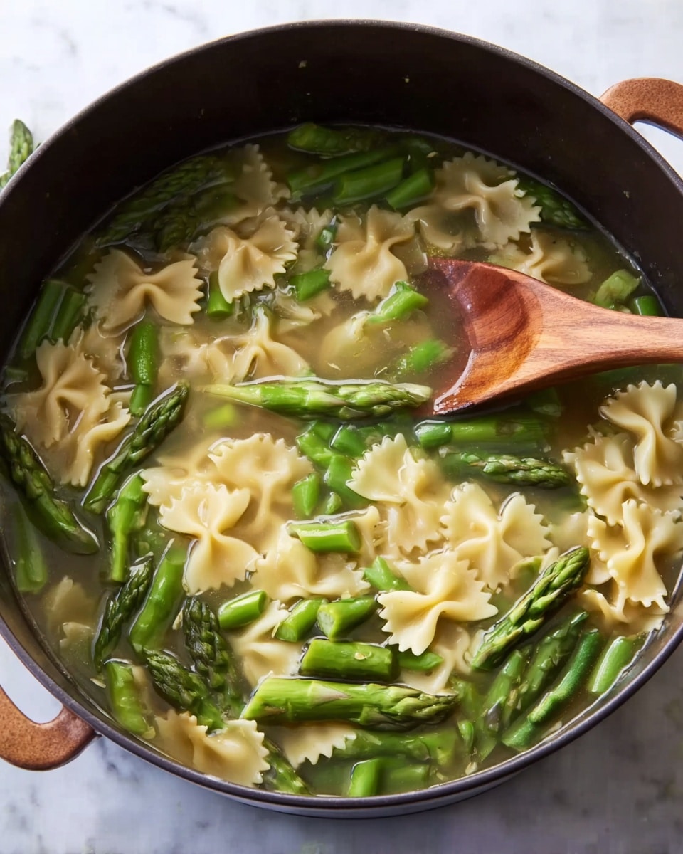 The image shows a large dark pot filled with a light broth containing three main layers: at the bottom are pale green soup liquid, the middle layer has short pieces of bright green asparagus evenly scattered, and the top layer has bow-tie shaped pasta in a light beige color floating among the asparagus. A wooden spoon with a smooth triangular head is partially submerged on the right side, stirring the soup. The pot sits on a white marbled surface with no visible plate or bowl. Photo taken with an iphone --ar 4:5 --v 7