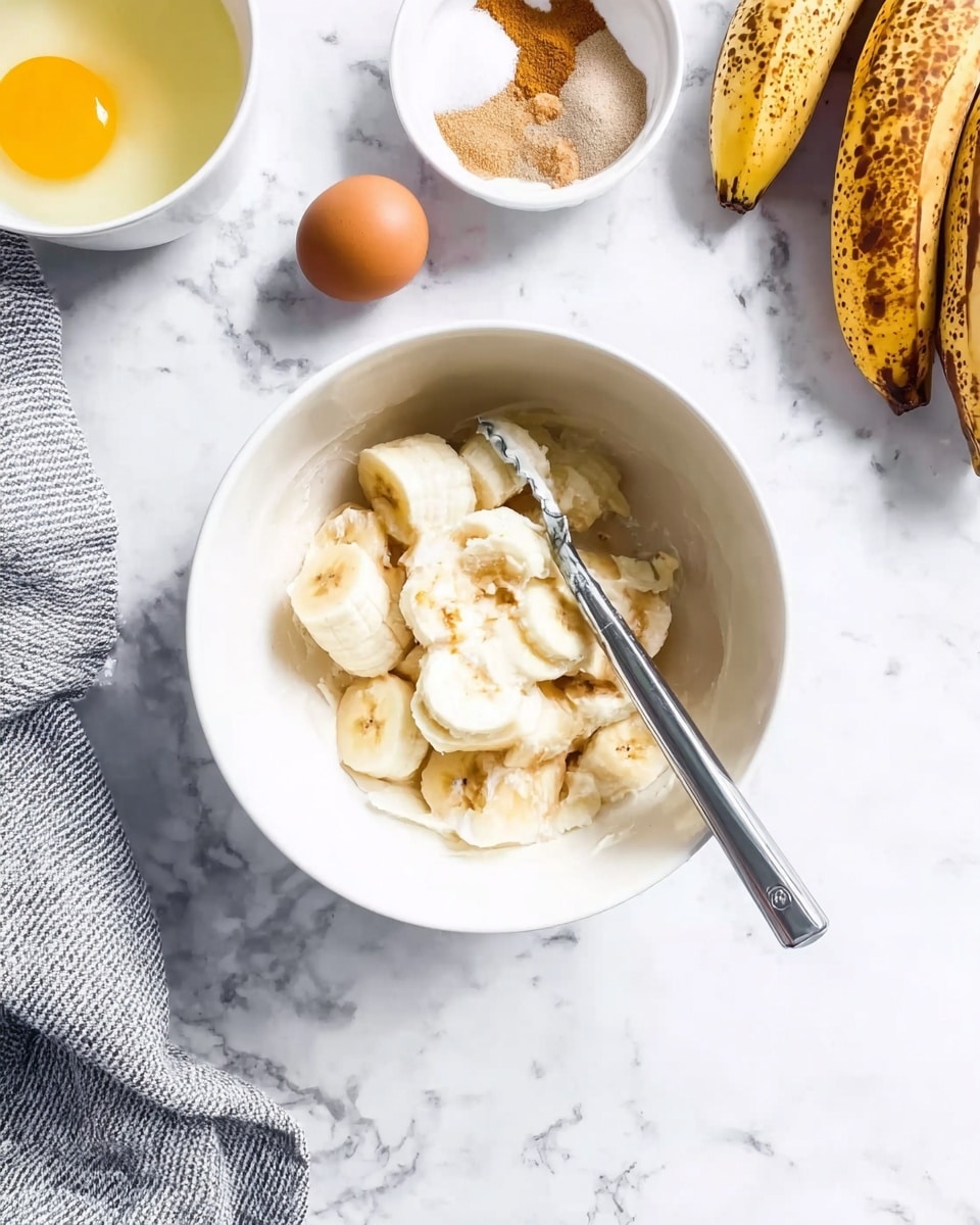 A white bowl sits on a white marbled surface, filled with peeled bananas being mashed by a silver masher, the bananas showing soft, creamy white flesh and small brown spots. To the left, a cracked raw egg is visible in a white bowl with bright yellow yolk, next to a single raw egg with brown shell. Above these, a small white bowl holds a mix of cinnamon powder and sugar, both in light brown and white colors. In the top right corner, several ripe plantains with yellow and brown skin rest on the marbled surface. A gray and white striped cloth is partly visible at the bottom left corner. Photo taken with an iphone --ar 4:5 --v 7