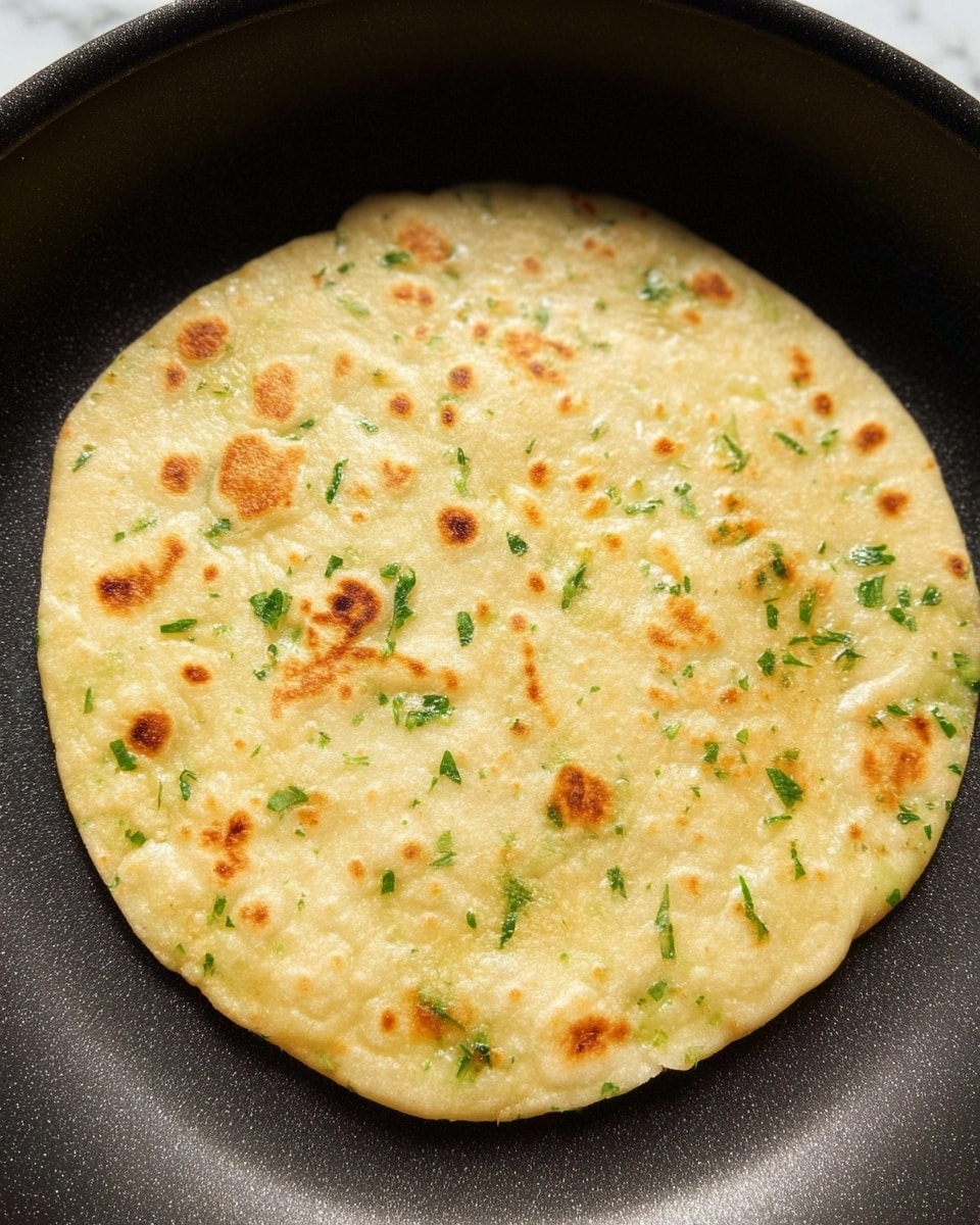 A close-up view of a single round flatbread cooking in a black pan, with small brown spots scattered across its light yellow surface. The flatbread has visible green herb pieces evenly spread through the dough, giving it a speckled look. The pan's texture is smooth and slightly shiny, showing the flatbread centered inside it. The flatbread looks soft and slightly thick with small air bubbles and gentle unevenness on its surface. The background is a white marbled texture photo taken with an iphone --ar 4:5 --v 7