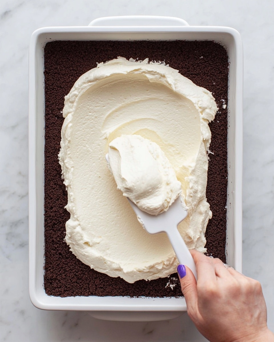 A white rectangular baking dish filled with a dark brown crumb layer, smooth and evenly pressed down at the bottom and sides. On top, there is a thick dollop of creamy white spread, soft and fluffy in texture, resting in the center with a white spatula partially covered in the spread, held by a woman's hand with purple polished nails, starting to spread it out. The dish sits on a white marbled surface. photo taken with an iphone --ar 4:5 --v 7