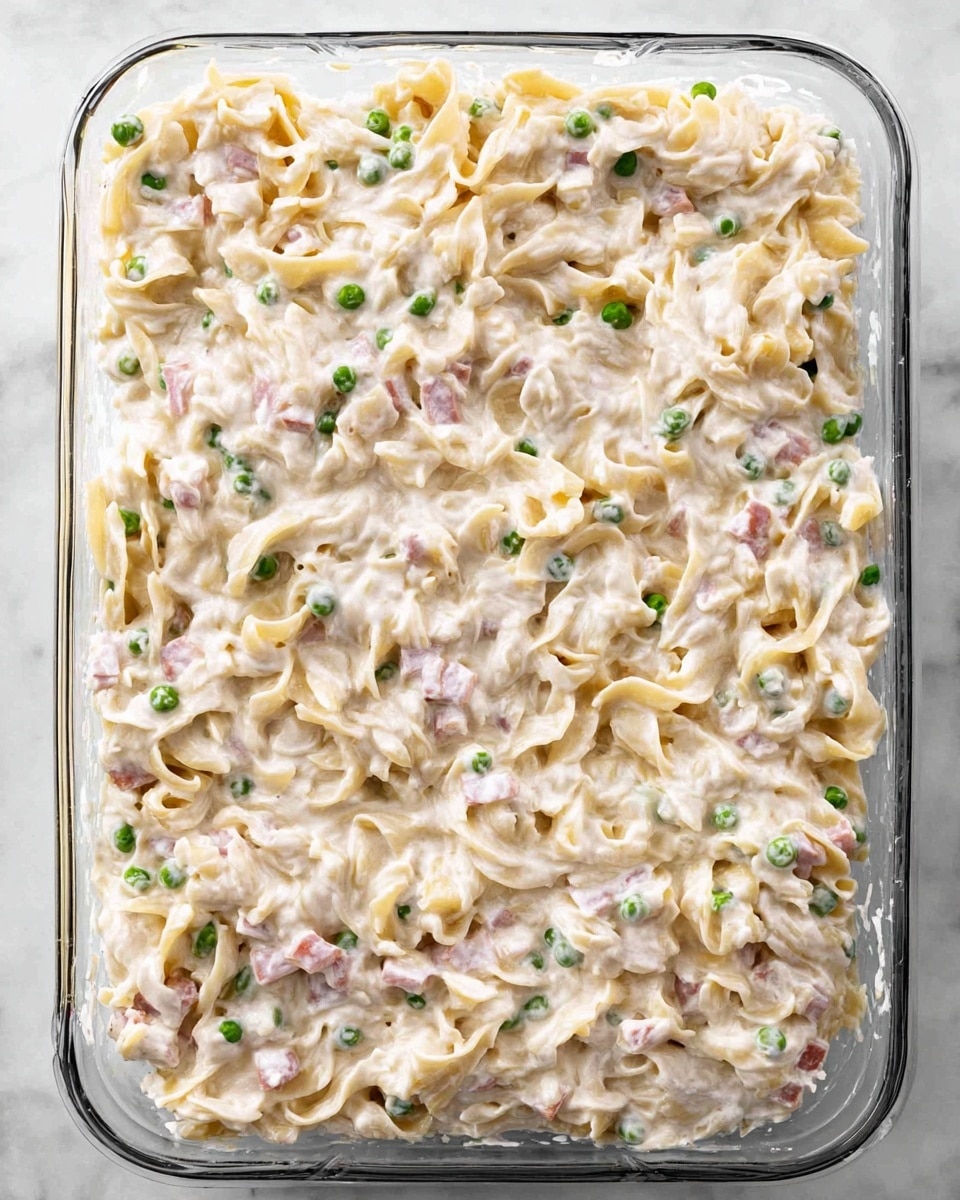 This image shows a rectangular glass baking dish filled with one thick layer of creamy pasta mixed with small green peas and bits of pink ham. The pasta appears to be egg noodles covered in a white sauce with a smooth, slightly lumpy texture. The dish sits on a white marbled surface, and there are no other items around it. photo taken with an iphone --ar 4:5 --v 7