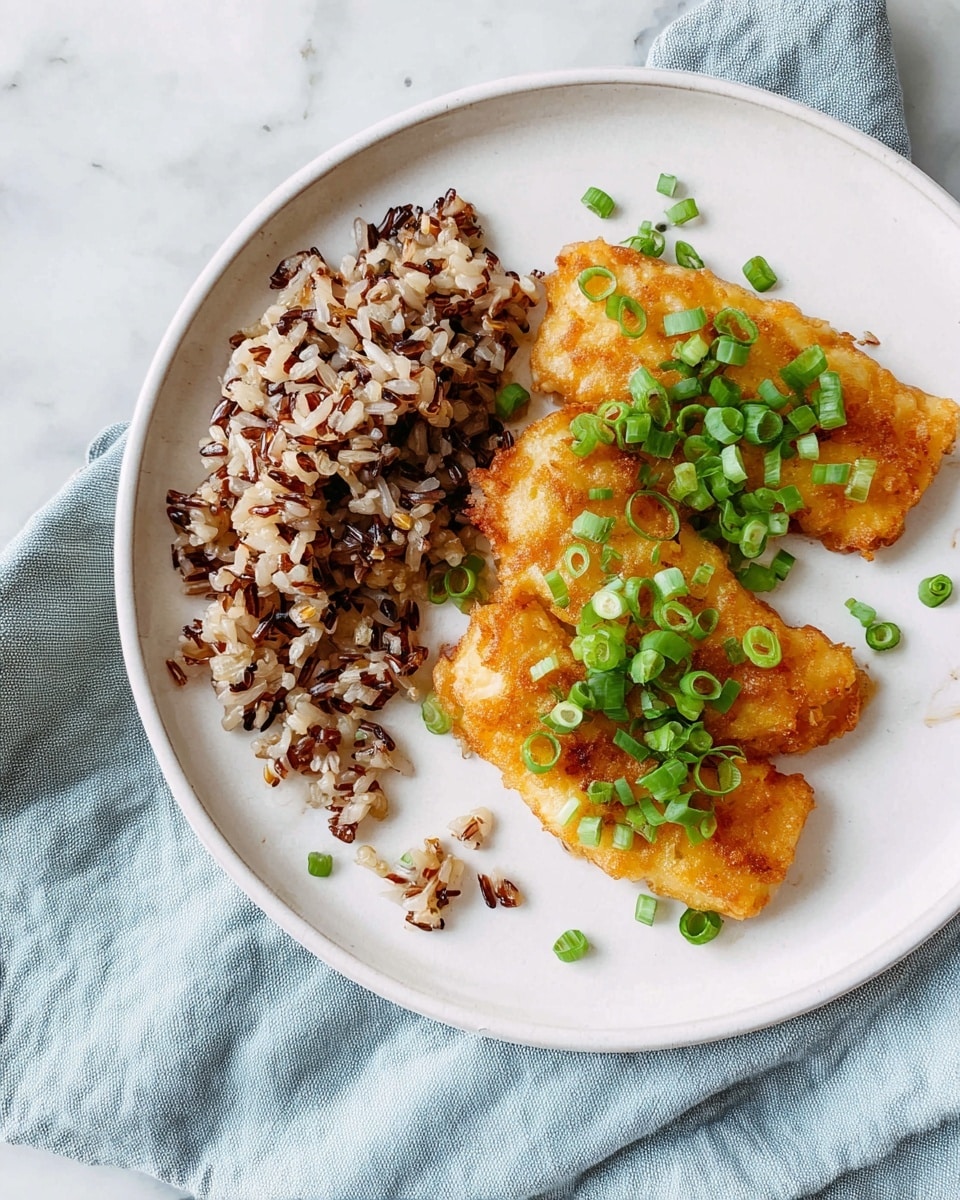 A white plate with two golden-brown fried pieces topped with bright green chopped scallions placed on the right side, and a clump of mixed brown and black rice positioned on the left side, all set on a light blue cloth over a white marbled surface. The fried pieces have a slightly crispy texture with small bits visible on top and around them. photo taken with an iphone --ar 4:5 --v 7