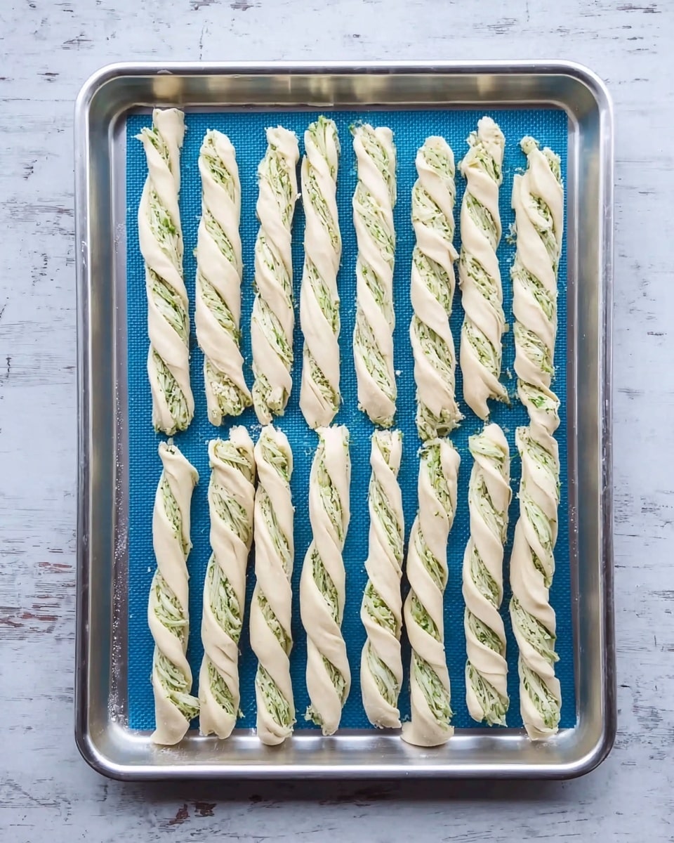 The image shows a silver baking tray with a blue silicone mat on top, holding fourteen uncooked twisted dough sticks. Each dough twist is pale, almost white, with green herb pieces mixed and spread inside the twisted layers. The twists are arranged in three rows with four in the top two rows and six in the bottom row. The background is a white marbled surface. photo taken with an iphone --ar 4:5 --v 7