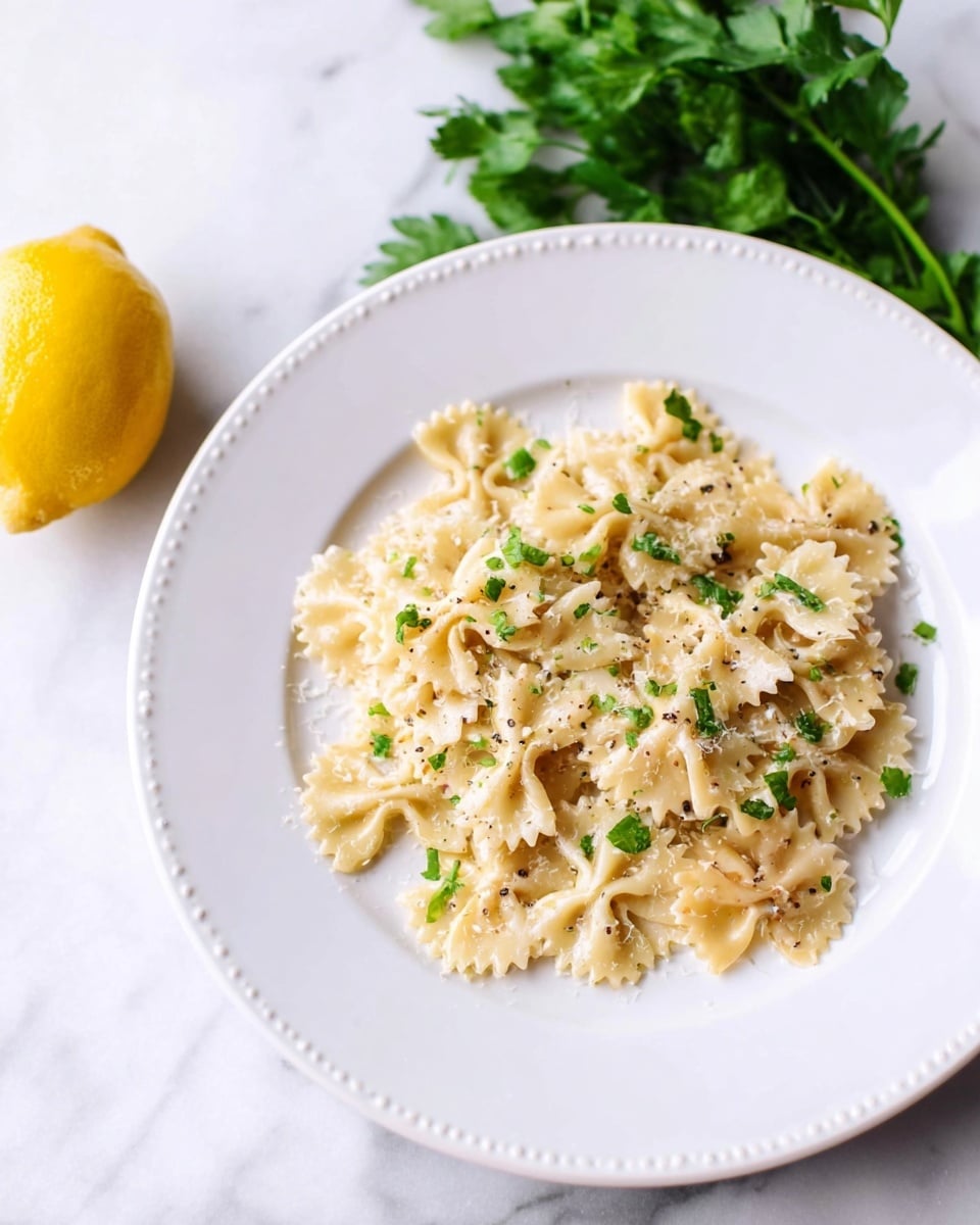A white plate with a small raised bead edge holds a single layer of bowtie pasta mixed with a light creamy sauce, sprinkled with small green herb pieces and black pepper specks. The sauce looks soft and slightly thick, covering the pasta unevenly. Around the plate, a bunch of fresh green parsley leaves sit at the top left and half a lemon with bright yellow skin rests at the bottom left on a white marbled surface. The scene is well-lit and simple, focusing on the gentle colors and textures of the pasta dish. photo taken with an iphone --ar 4:5 --v 7