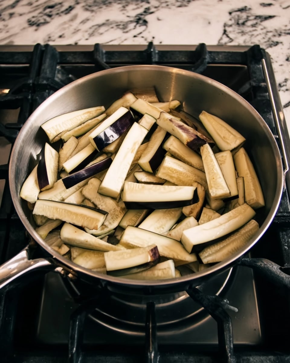 A shiny stainless steel pan sits on a gas stove with its burner on, filled with many pieces of eggplant. The eggplant pieces are cut into thick, long wedges, mostly light cream in color with dark purple skin edges showing. They fill the pan in a loose, uneven layer, some resting on top of others. The stove and countertop under the pan have a granite look, but the visible part of the image background has been changed to a white marbled texture. photo taken with an iphone --ar 4:5 --v 7