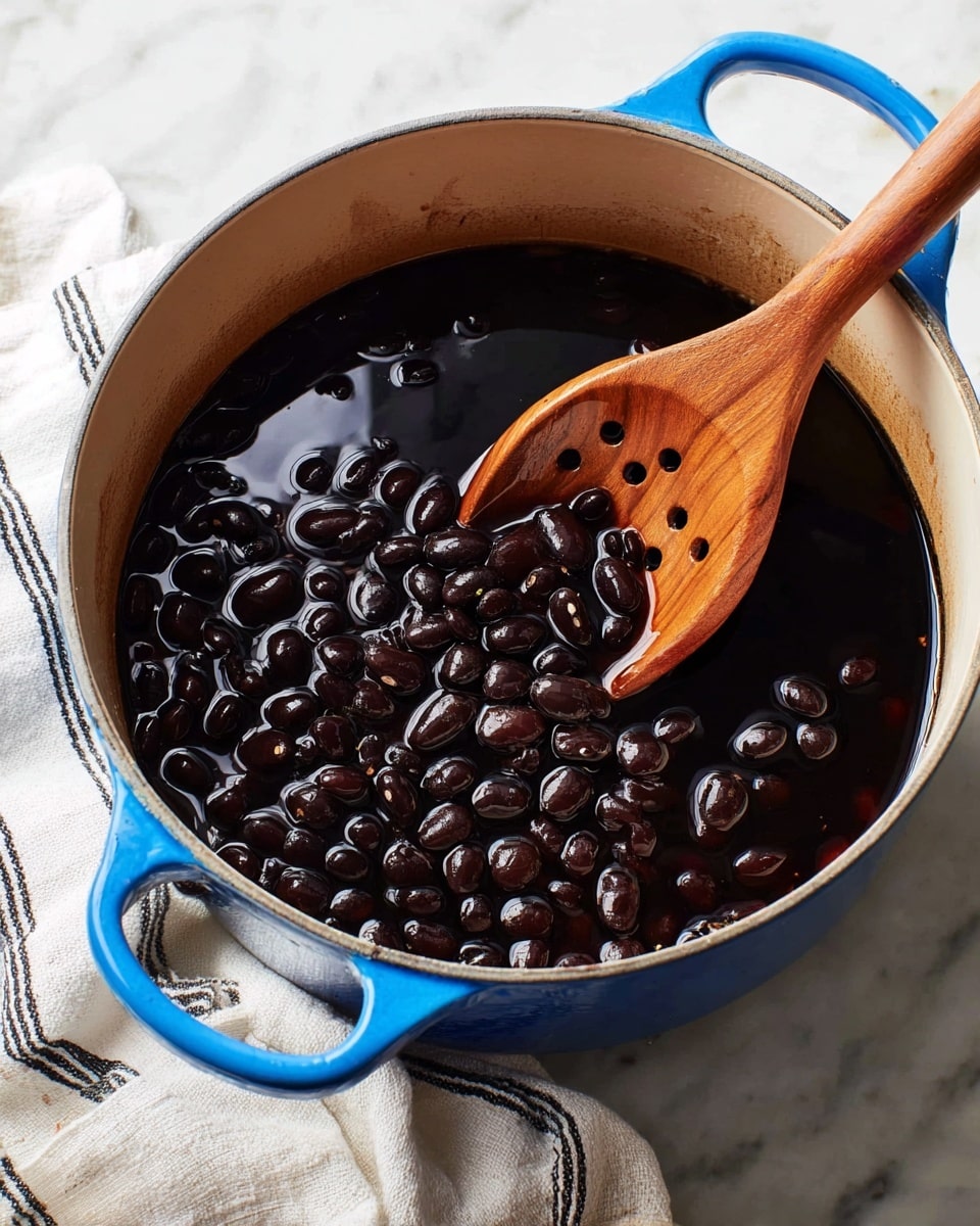 A blue pot filled with shiny black beans submerged in dark liquid, showing a thick layer of beans on top with some floating in the middle. A wooden spoon with holes is partially inside the pot, resting on the beans and liquid. The pot sits on a white marbled surface with a white cloth featuring black stripes underneath the pot’s handles. The lighting highlights the glossiness of the beans and the smooth texture of the wooden spoon. photo taken with an iphone --ar 4:5 --v 7