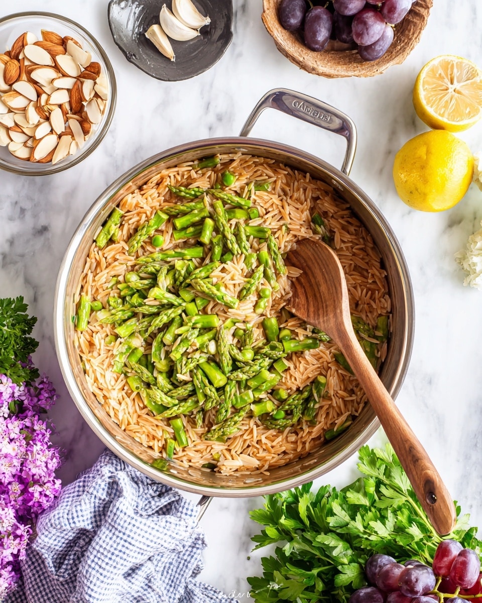 A metal pan sits on a white marbled surface, holding two main layers: a base of light brown orzo pasta that fills most of the pan and a top layer of chopped green asparagus pieces covering about half of the pasta. A wooden spoon with a smooth texture rests inside the pan, partially mixing the layers. Around the pan, there are small bowls with ingredients: one bowl with light beige slivered almonds, a dark small plate with garlic cloves, a halved lemon showing its bright yellow interior, grapes in a clear bowl, and fresh green parsley alongside purple flowers. A white and blue checkered cloth is placed near the pan. Photo taken with an iphone --ar 4:5 --v 7
