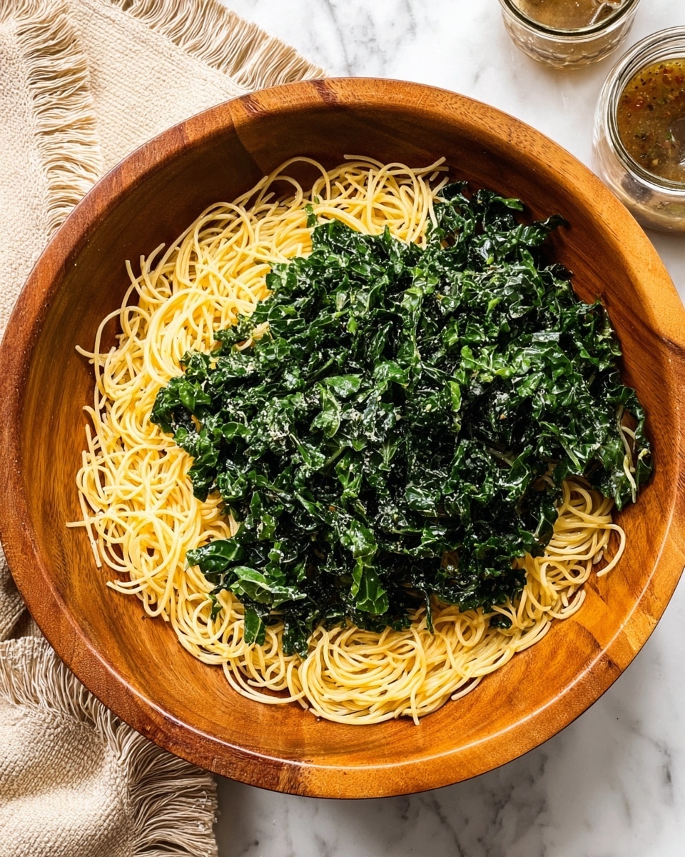 The image shows a large wooden bowl filled with two main layers of food. The bottom layer is light yellow thin spaghetti noodles, loosely spread across the bowl. On top is a thick layer of dark green, chopped cooked kale that covers most of the noodles but allows some of the yellow strands to peek through, giving a mix of colors and textures. The bowl sits on a white marbled surface with a beige fringed cloth partially visible in the top left corner and a glass jar of sauce or dressing in the top right. photo taken with an iphone --ar 4:5 --v 7