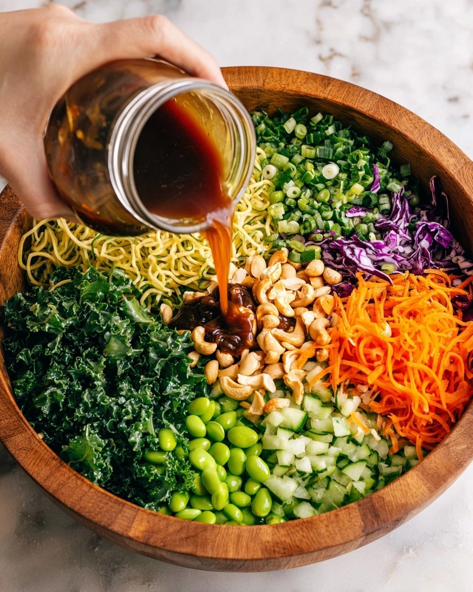 A large wooden bowl filled with many fresh ingredients arranged in separate layers: bright green chopped cilantro, dark green chopped kale, light yellow cooked noodles, dark purple shredded cabbage, light green sliced cucumber, dark green chopped herbs, chopped green onions, orange shredded carrots, bright green edamame beans, and light brown whole cashews. A woman's hand is pouring a dark brown sauce from a clear glass jar into the center of the bowl. The background surface is a white marbled texture. Photo taken with an iphone --ar 4:5 --v 7