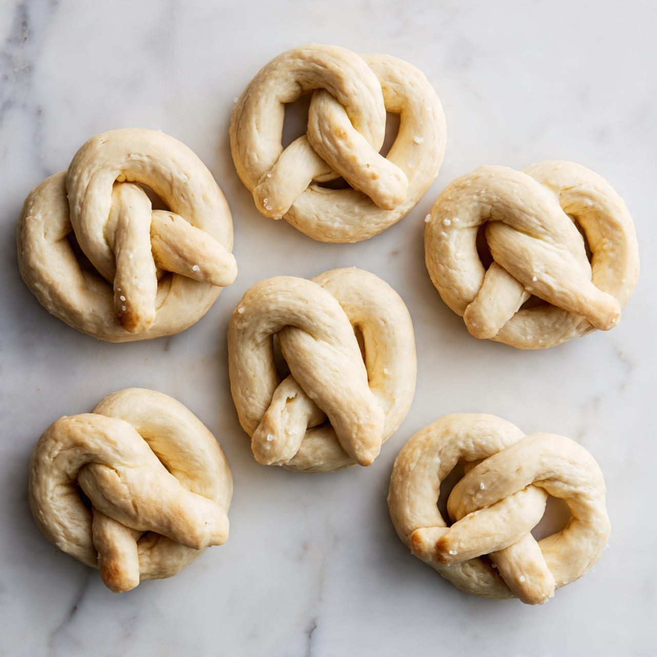 The image shows six raw pretzels placed on a white marbled surface. Each pretzel is made from a single piece of pale, smooth dough twisted into the classic knot shape with two thick loops on top and the ends crossing over in the center. The dough texture shows slight creases and folds where it twists. The pretzels are evenly spaced over the surface, with varied but similar shapes and sizes, all uncooked and unbaked, maintaining a soft dough look. photo taken with an iphone --ar 4:5 --v 7