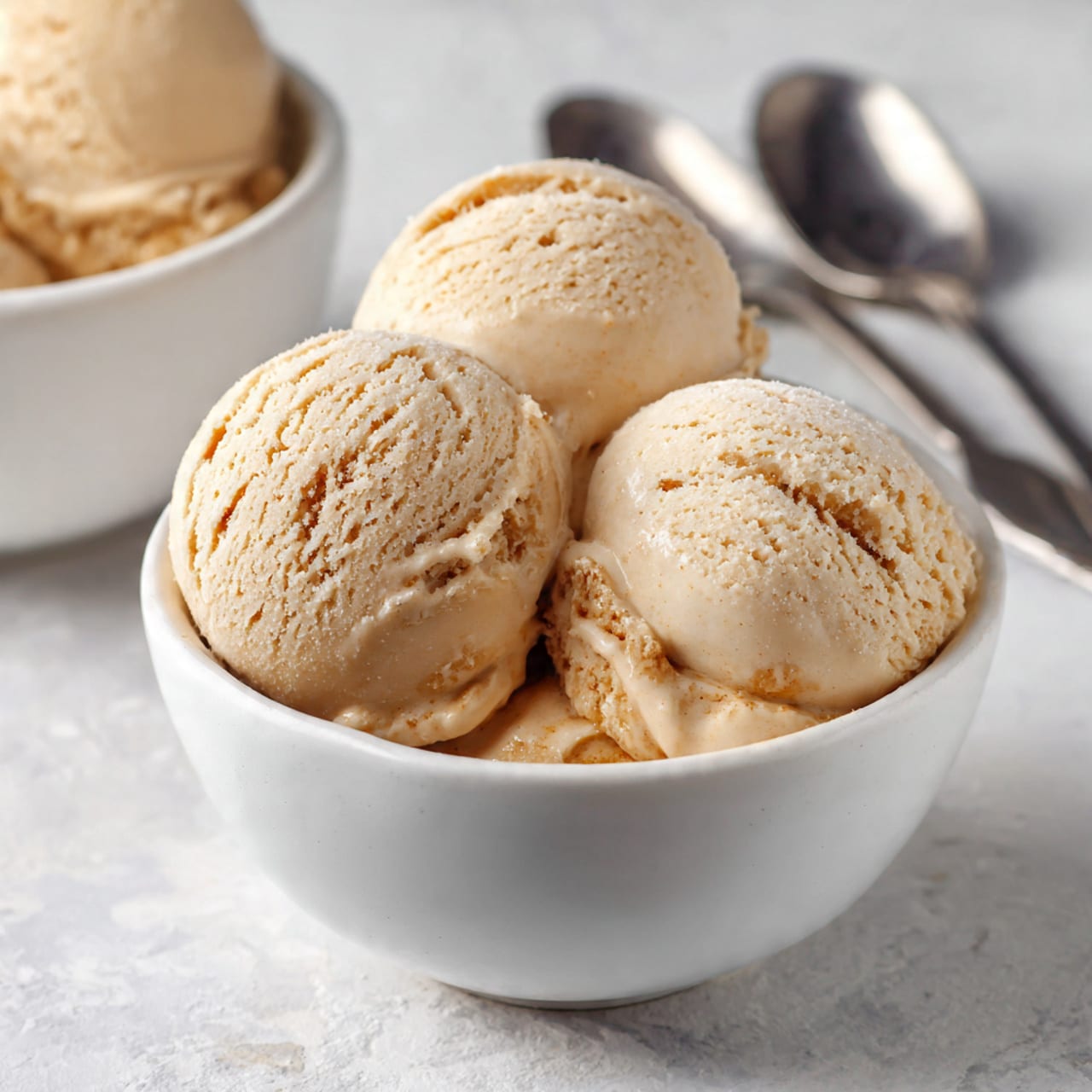 Three smooth light brown ice cream scoops sit close together in a white bowl in the front, showing some soft texture and small white swirls. Behind it, a second white bowl holds more similar scoops, slightly blurred. To the upper right, two shiny silver spoons lay side by side on a white marbled surface. The scene is bright and clean, focused on the creamy ice cream texture and soft colors, photo taken with an iphone --ar 4:5 --v 7