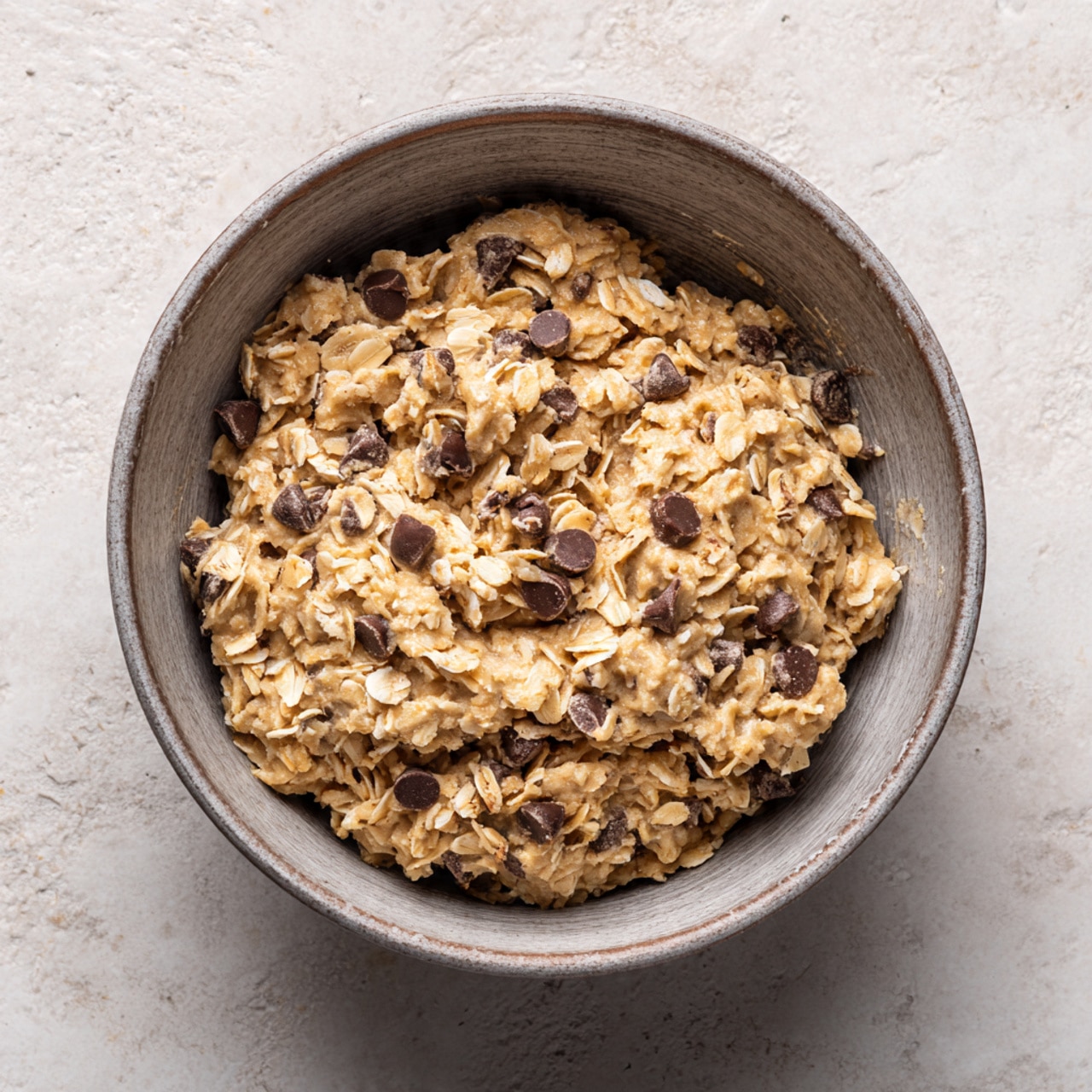 A close-up view of an unbaked oatmeal chocolate chip cookie dough inside a gray metal mixing bowl. The dough has a rough texture with visible rolled oats and scattered small dark brown chocolate chips throughout. The dough is packed into the bowl, showing a light brown color mixed with creamy oat flakes and darker chocolate bits. The background is a white marbled texture. photo taken with an iphone --ar 4:5 --v 7