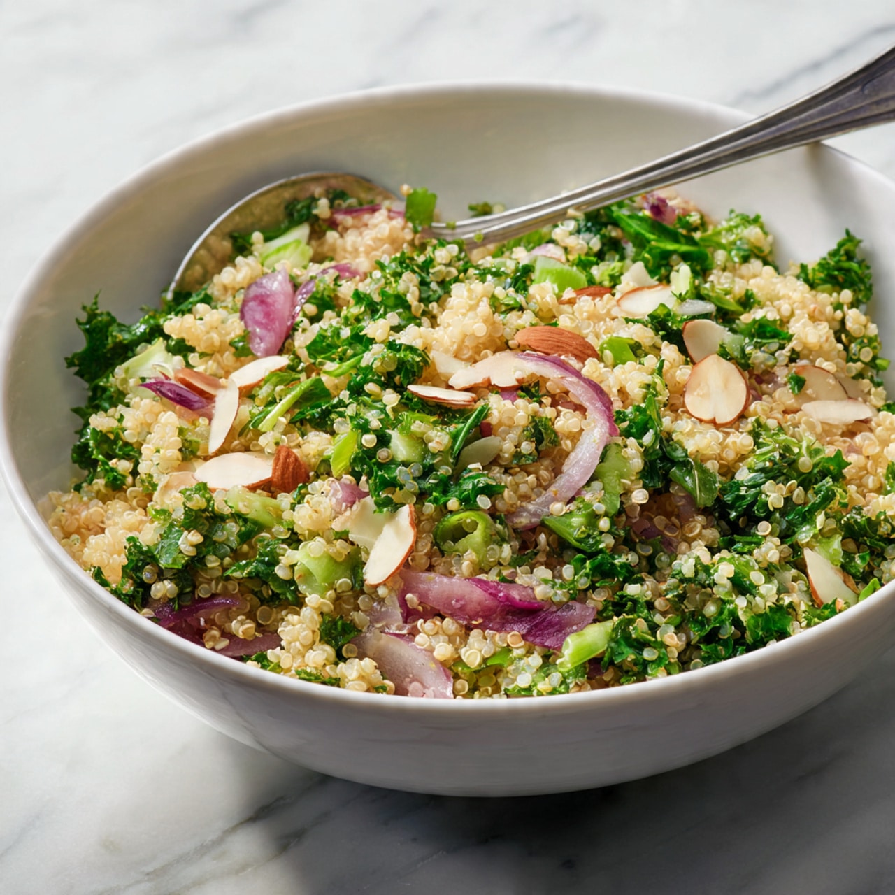 A white bowl filled with a grain salad made of small, light beige quinoa grains as the base layer. Mixed inside are chopped bright green leafy vegetables scattered evenly, along with diced pinkish-purple onion pieces. There are also thin, light brown toasted almond slices spread on top. A silver fork is resting inside the bowl near the upper edge. The bowl is placed on a white marbled surface. photo taken with an iphone --ar 4:5 --v 7