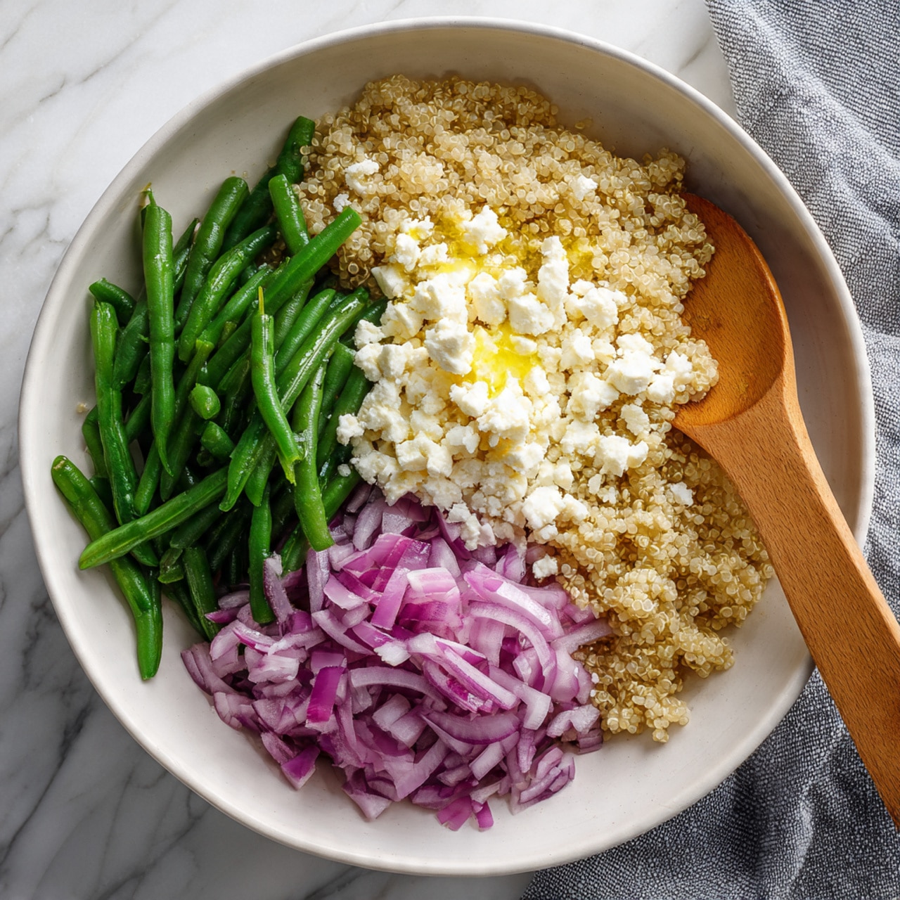 The image shows a white bowl filled with cooked quinoa as the base layer, topped with three separate piles: one of chopped green beans with a fresh, firm texture on the top left, another of small white cheese crumbles with a soft, crumbly texture on the top right drizzled lightly with a yellow liquid, and a third of finely chopped red onions with a crisp texture on the bottom left. A wooden spoon rests inside the bowl on the right side. The bowl is placed on a white marbled surface with a grey cloth partly visible on the bottom right photo taken with an iphone --ar 4:5 --v 7