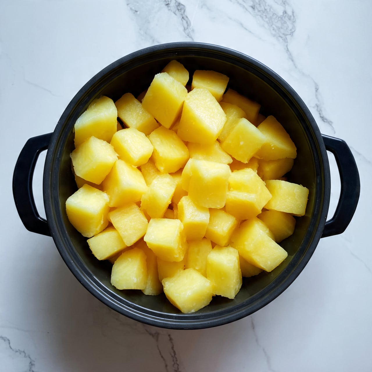 A black pot filled to the top with small, evenly-sized cubes of yellow boiled potatoes is shown from above. The potato cubes have a soft and smooth texture with gentle edges. The pot has two black handles on either side and is placed on a white marbled surface. Photo taken with an iphone --ar 4:5 --v 7