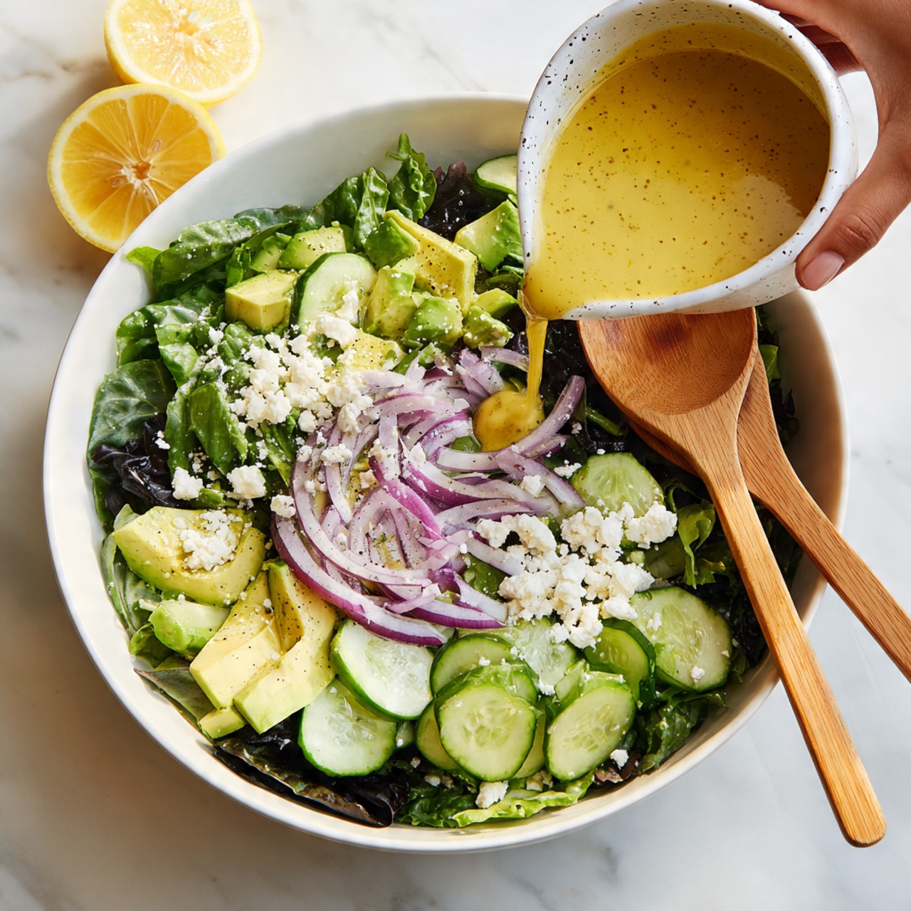 A white bowl holds a fresh salad with three main layers: the bottom layer is dark green leafy vegetables, the middle layer features sliced light green cucumbers and chunks of light yellow avocado, and the top layer is made of thin purple onion slices scattered over the salad along with small white cheese crumbles. A woman’s hand is pouring a yellow dressing from a white speckled container onto the salad, while two wooden spoons rest inside the bowl. The bowl sits on a white marbled surface, and part of a lemon wedge is visible in the background. Photo taken with an iphone --ar 4:5 --v 7