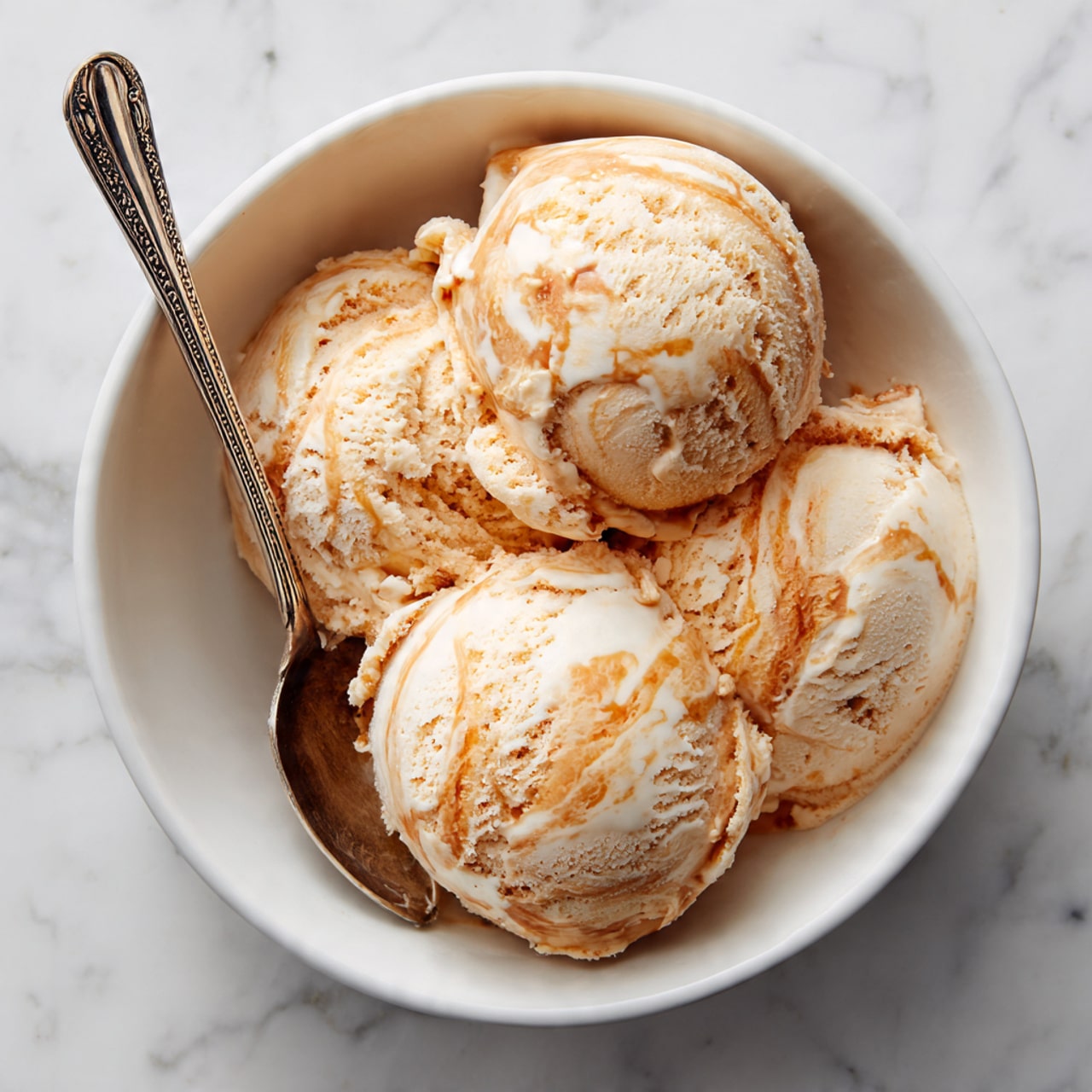 The image shows a white bowl with three scoops of light brown ice cream, each scoop smooth with slight texture and some creamy white swirls and patches. The scoops are close together inside the bowl. A silver spoon with a detailed handle is placed on the left side of the bowl, resting partly in the ice cream. The bowl sits on a surface with a white marbled texture. photo taken with an iphone --ar 4:5 --v 7