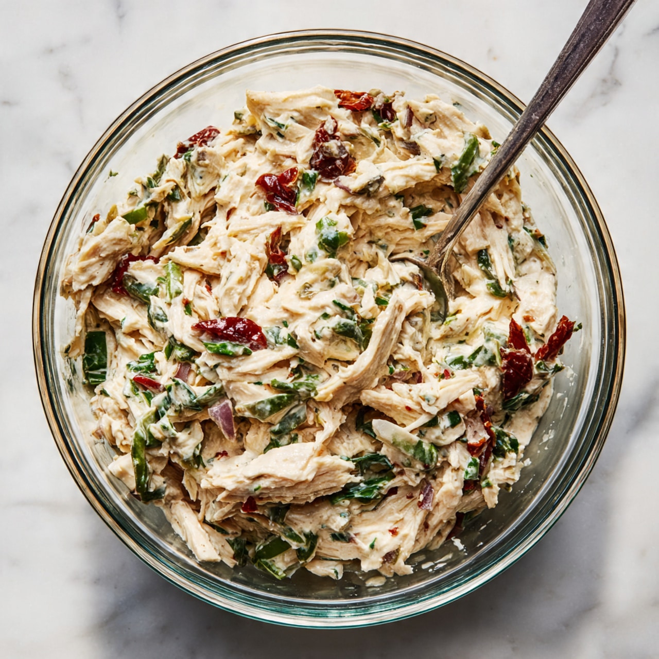 A clear glass bowl filled with a creamy mixture made of shredded light beige chicken combined with small pieces of bright green celery, dark green spinach or herbs, tiny diced purple onions, and reddish-brown sun-dried tomatoes. The mix shows a soft, thick texture with visible bits of each ingredient evenly spread throughout. The bowl sits on a white marbled surface, and a metal spoon stands on the right side inside the bowl. The photo taken with an iphone --ar 4:5 --v 7