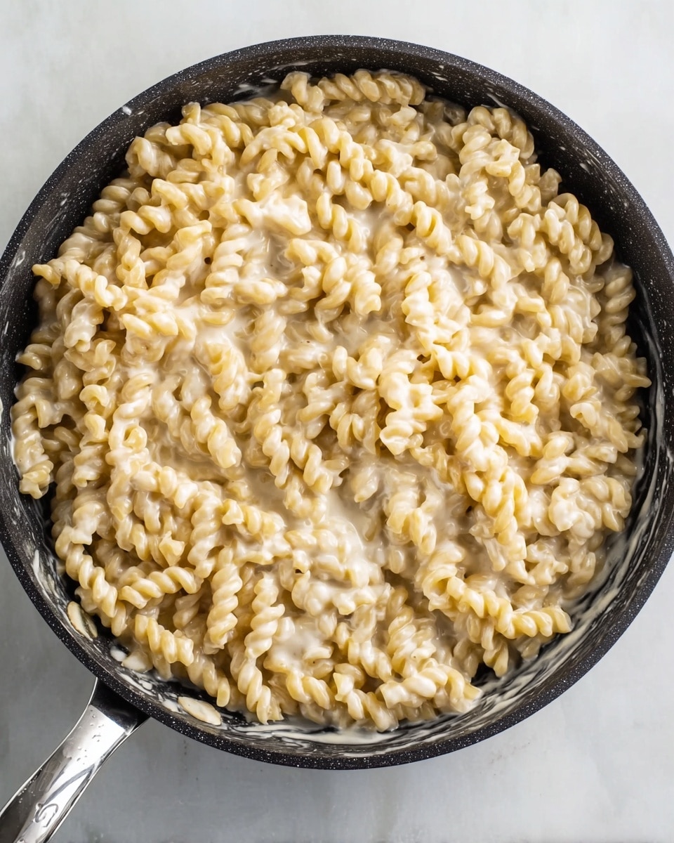 A black pan filled with a single thick layer of small, twisted, light golden pasta pieces coated in a creamy white sauce. The creamy sauce appears smooth and slightly melted, covering most of the pasta evenly with some sauce pooling at the edges of the pan. The pan has a shiny silver handle, and it sits on a white marbled textured surface. Photo taken with an iphone --ar 4:5 --v 7