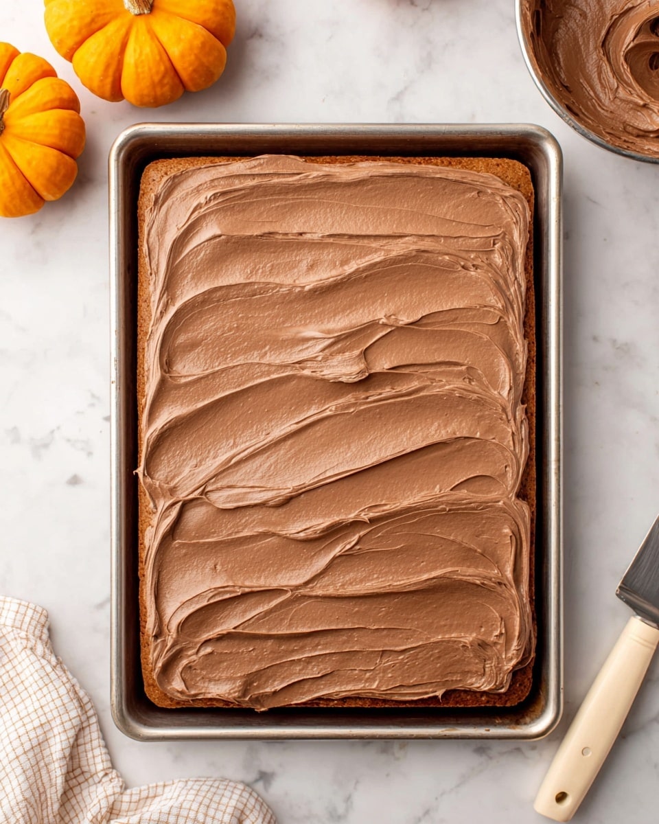 The image shows a single rectangular layer of light brown cake in a baking tray, topped with a thick, smooth layer of milk chocolate frosting spread evenly with soft, wavy patterns. The frosting covers the entire surface of the cake with some edges gently spilling over the tray. The tray sits on a white marbled surface with two small orange pumpkins in the top left corner, a metal bowl with more chocolate frosting in the top right, the tip of a beige knife handle on the bottom left, and a white and beige checkered cloth partially visible on the lower right side. Photo taken with an iphone --ar 4:5 --v 7