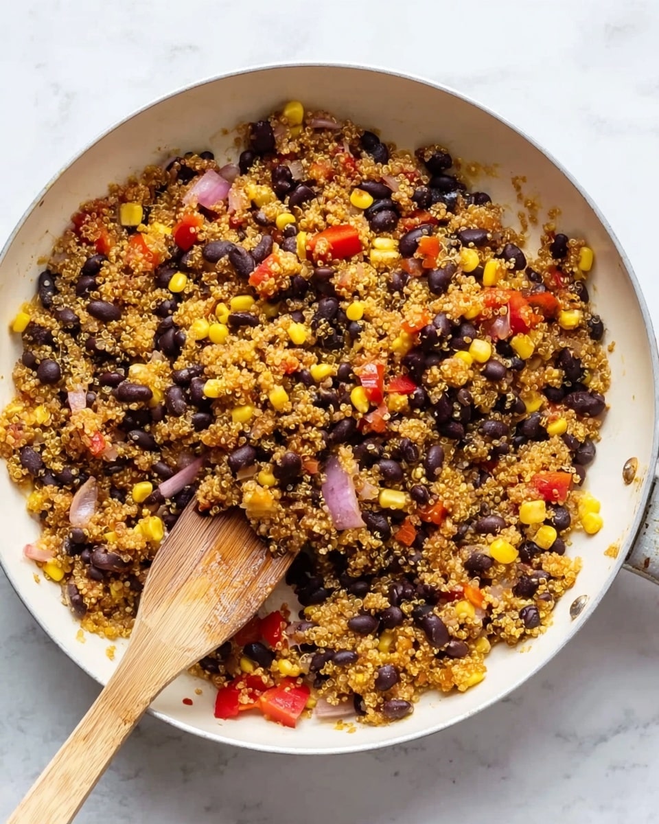 A white frying pan filled with a mix of cooked quinoa, black beans, corn kernels, and small pieces of red bell pepper and red onion. The ingredients are evenly spread, showing a colorful combination of yellow, black, red, and light purple with a slightly grainy and soft texture. A wooden spatula is resting on the right side inside the pan, partially covered by the food, with its smooth wooden surface visible. The pan is set on a white marbled surface. photo taken with an iphone --ar 4:5 --v 7