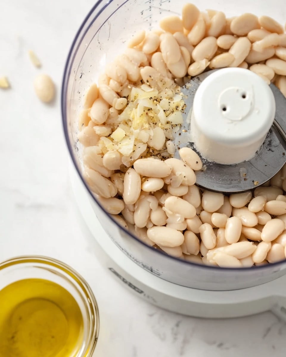 A close-up image of a white food processor with a clear bowl filled with light beige white beans and small pieces of pale yellow garlic, mixed with tiny specks of black pepper. The food processor's blade is visible in the center, surrounded by the beans. To the left foreground, a clear glass cup with golden yellow olive oil is partially visible on a white marbled surface. The background is also a white marbled texture. The setting looks clean and bright, focusing on the raw ingredients ready to be blended photo taken with an iphone --ar 4:5 --v 7