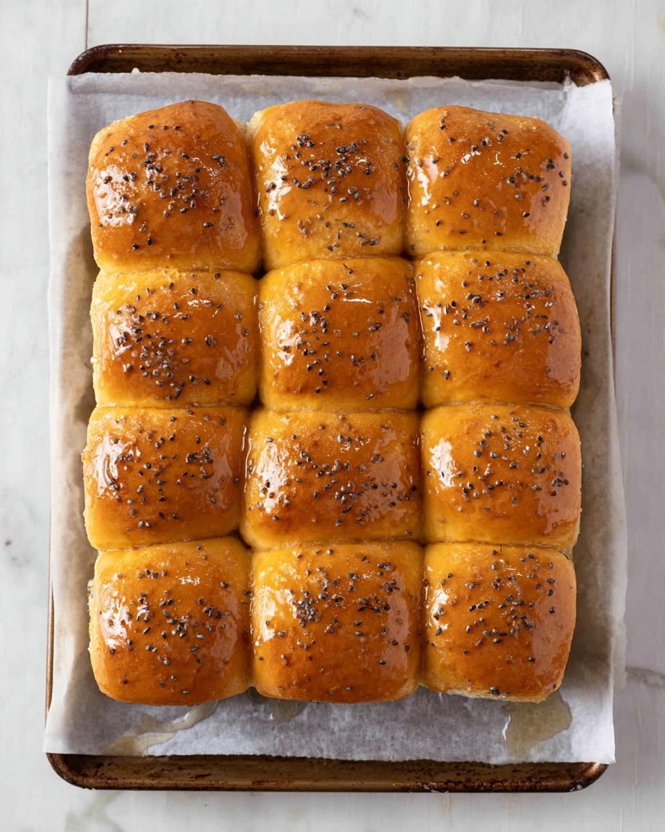 A tray of 16 soft, golden brown dinner rolls arranged in a 4 by 4 grid on white parchment paper, placed on a baking sheet with a slightly darkened edge. Each roll is shiny and smooth on top, covered with a light coating of glaze that reflects light, and sprinkled with small dark seeds that add texture. The rolls are closely packed, slightly touching each other with visible soft layers underneath the shiny top crust. The whole tray is set on a white marbled surface, showing a simple and clean background. photo taken with an iphone --ar 4:5 --v 7