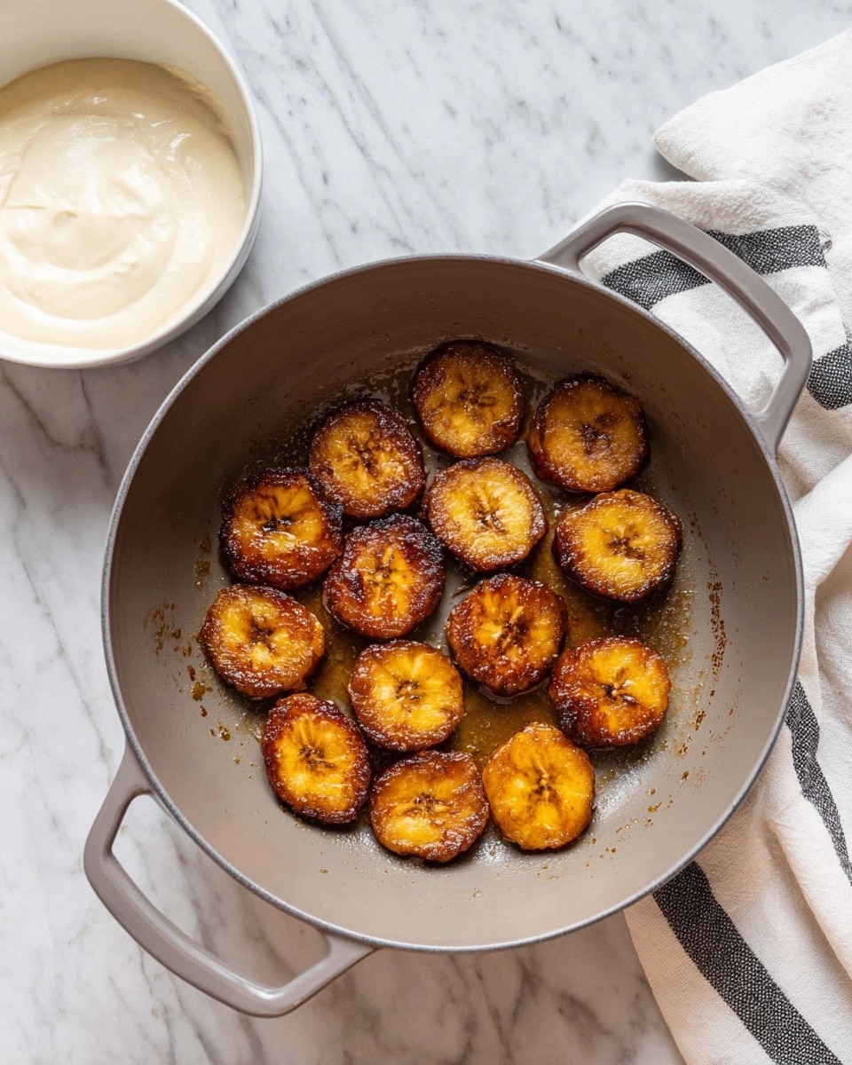A gray pan with two handles is placed on a white marbled surface, inside it are 13 fried plantain slices arranged around the center. The plantains are golden brown with a slightly shiny, caramelized texture from cooking oil. To the left of the pan, there is a white bowl filled with thick, creamy white sauce. On the right side, there is a folded white cloth with black stripes resting on the surface. The lighting highlights the shiny caramelized edges of the plantains and the smooth texture of the sauce. photo taken with an iphone --ar 4:5 --v 7