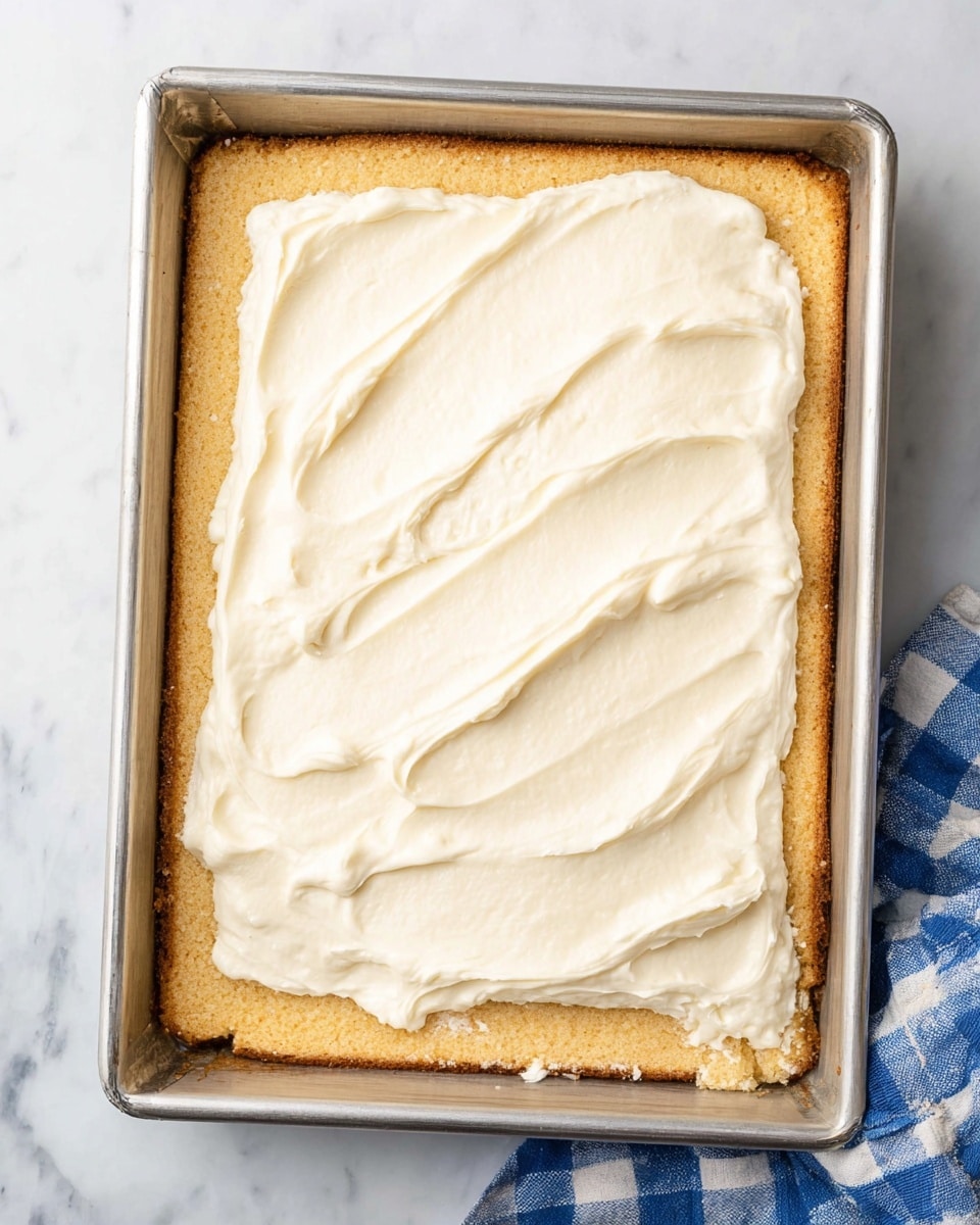 A rectangular metal baking pan holds a two-layer dessert. The bottom layer is a light golden, crumbly crust that covers the entire pan evenly, with slightly rough edges where it meets the pan sides. On top, creamy white frosting is being spread, covering about half the crust with a smooth, glossy texture and soft swirling patterns. The pan sits on a white marbled surface, and a blue and white checkered cloth is partially visible at the bottom right corner. photo taken with an iphone --ar 4:5 --v 7