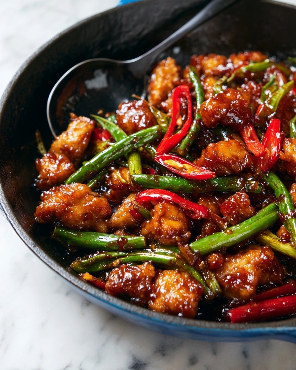 This close-up image shows a stir-fry dish with layers of small, browned pieces of meat mixed with bright green, slightly crunchy green beans and thin slices of red chili peppers scattered throughout. The food looks shiny and coated with a rich, dark sauce, with the meat having a crispy texture with some caramelized edges. A wooden spatula is partially visible on the left side, slightly lifting some of the ingredients. The dish is served in a dark pan, with the background being a white marbled surface. photo taken with an iphone --ar 4:5 --v 7