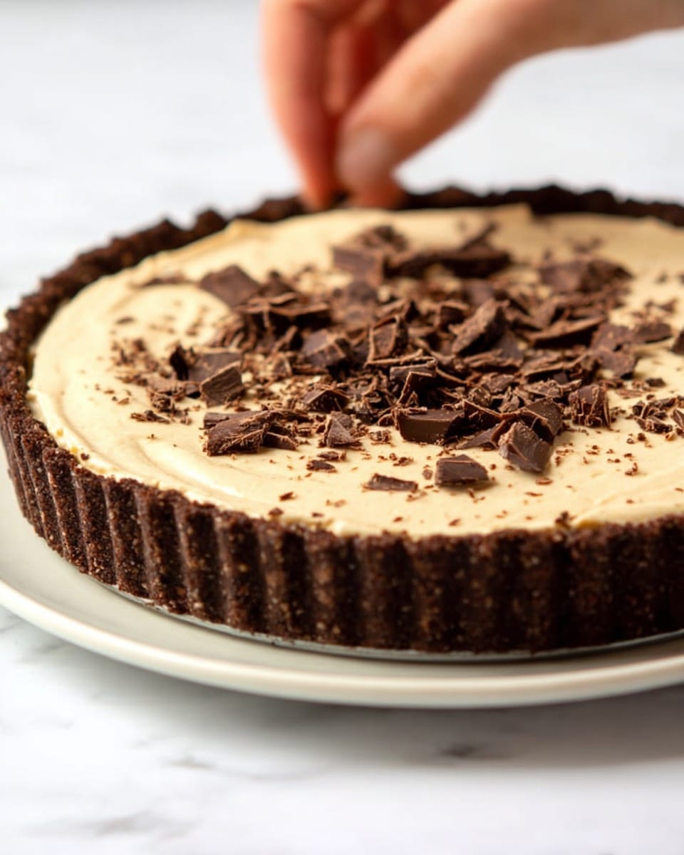 A round tart with a dark brown, crumbly crust forms the base layer, holding a smooth, light beige creamy filling in the middle. The creamy layer is thick and evenly spread, slightly rising above the crust edges. Small chunks and shavings of dark chocolate are being sprinkled on top by a woman's hand, adding texture and contrast to the smooth cream. The tart sits on a white plate, placed on a white marbled surface. The overall look is rich and inviting with a balance of dark and light colors. photo taken with an iphone --ar 4:5 --v 7