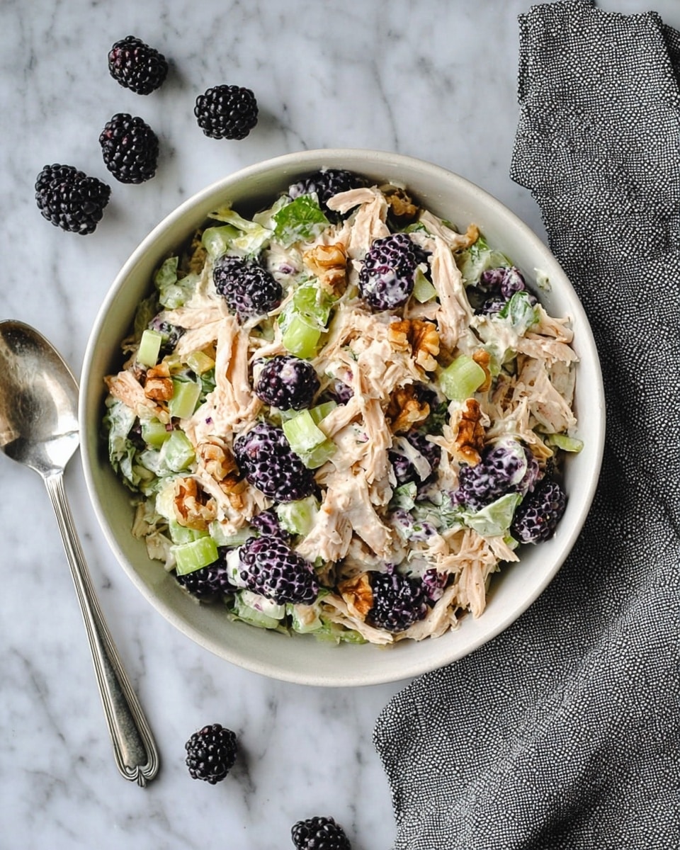 A bowl filled with a mixed salad containing shredded light beige chicken, small green celery pieces, dark purple blackberries, and small brown walnut pieces all coated with a creamy white sauce. The bowl is white and placed on a white marbled surface with three blackberries and a silver spoon near it, along with a grey textured cloth on the right side. photo taken with an iphone --ar 4:5 --v 7