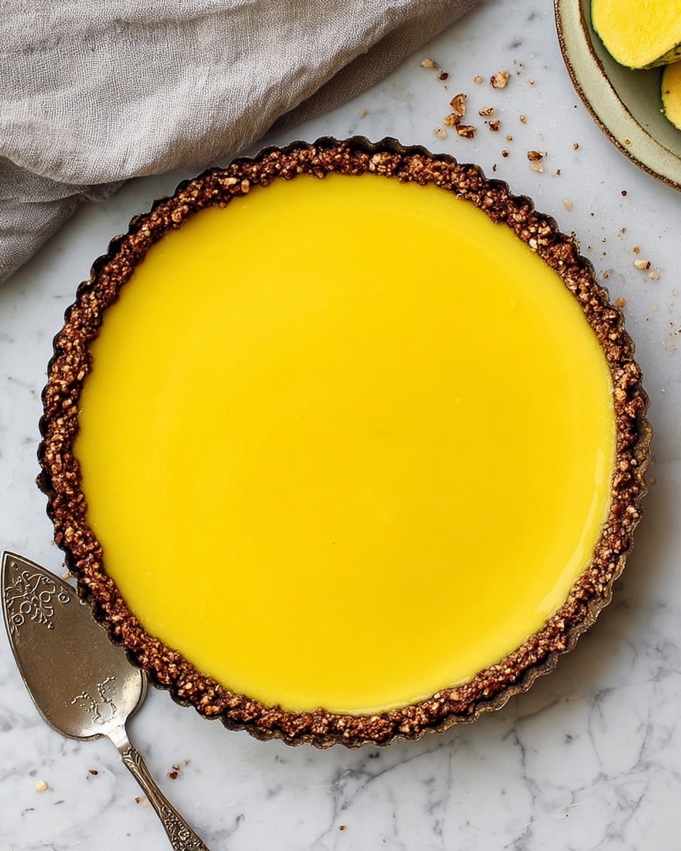 A round tart with a smooth, bright yellow top layer fills the tart shell evenly, with a thin, dark brown crust that has a rough texture with small nut pieces visible all around the edge. The tart sits on a white marbled surface with crumbs scattered nearby. A silver serving spatula with a decorative handle rests to the lower left side of the tart, and a piece of grey fabric is folded in the upper left corner. The top right corner shows the edge of a yellow fruit or vegetable slice. Photo taken with an iphone --ar 4:5 --v 7