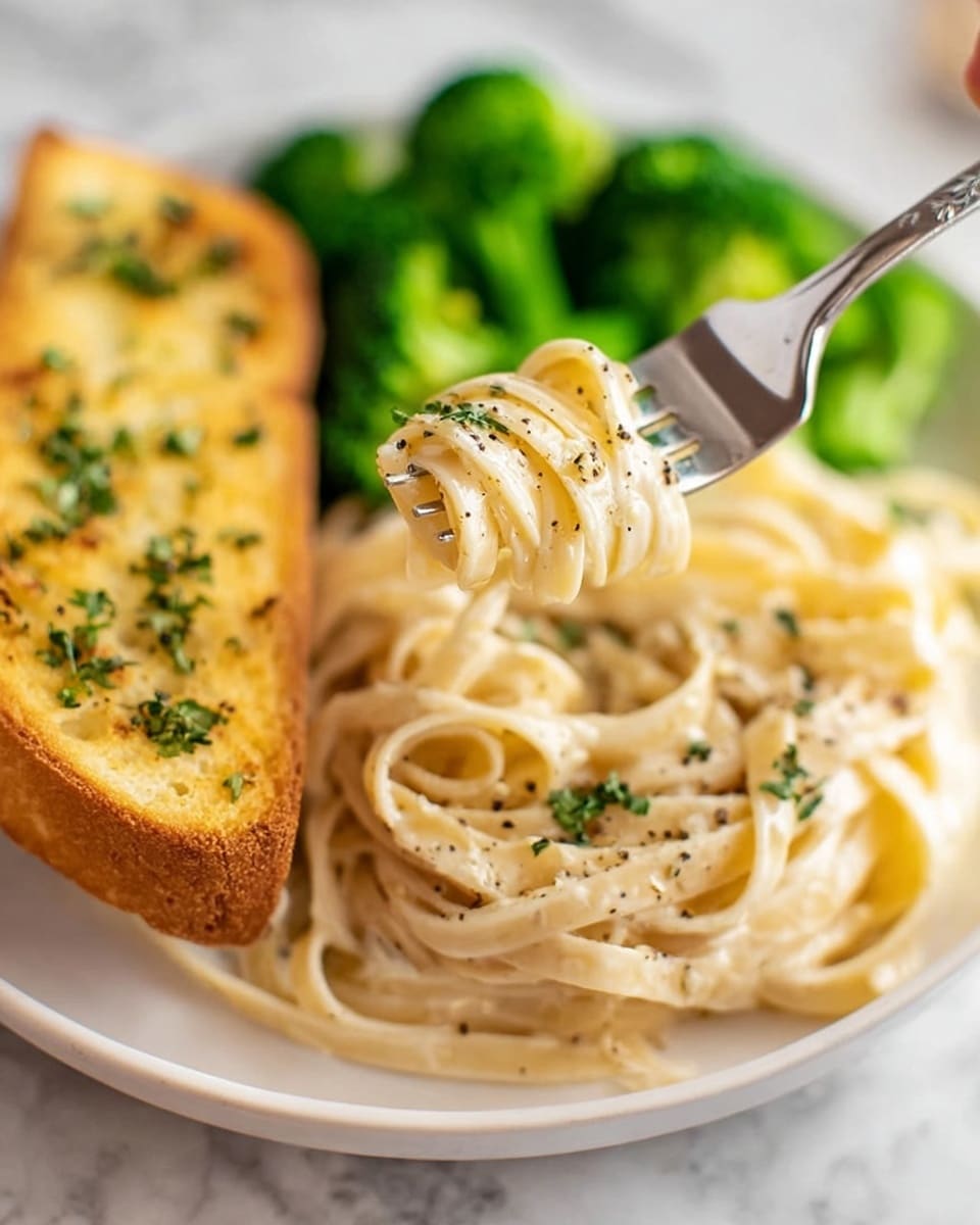 A white plate holds three main parts: on the left, a golden piece of garlic bread with bits of herbs on top; in the middle, a serving of creamy fettuccine pasta with a smooth white sauce, sprinkled with small green herb pieces and black pepper; at the back, bright green steamed broccoli florets add color and texture. The plate sits on a white marbled surface. photo taken with an iphone --ar 4:5 --v 7