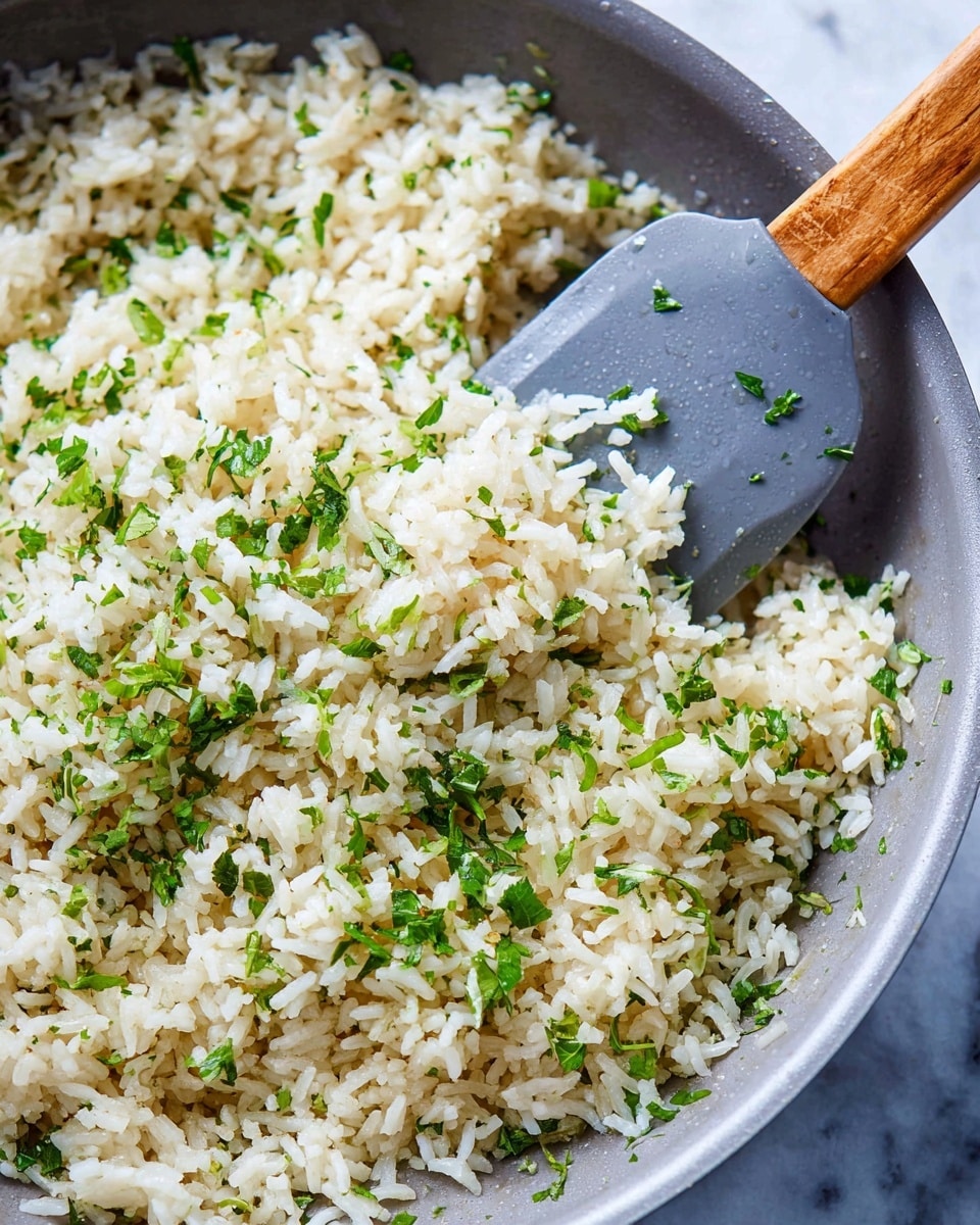 A close-up of a light gray pan filled with white rice mixed with chopped green herbs scattered evenly throughout. A gray spatula with a wooden handle is stirring the rice on the left side of the pan, lifting some rice showing its fluffy texture. The rice grains are plump and moist with a slight shine, and the herbs add small bright green spots that contrast with the pale color of the rice. The background surface has a white marbled texture. Photo taken with an iphone --ar 4:5 --v 7