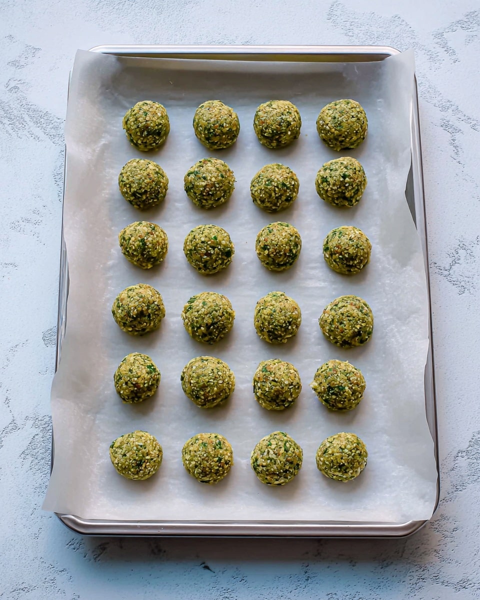 The image shows a rectangular baking tray lined with white parchment paper, holding 24 small, round green falafel balls arranged in four rows of six each. The falafel balls have a coarse texture with visible bits of herbs and chickpeas, and they sit evenly spaced on the paper. The tray is set on a surface with a white marbled texture, and the angle is from above, capturing the entire tray in clear light. photo taken with an iphone --ar 4:5 --v 7