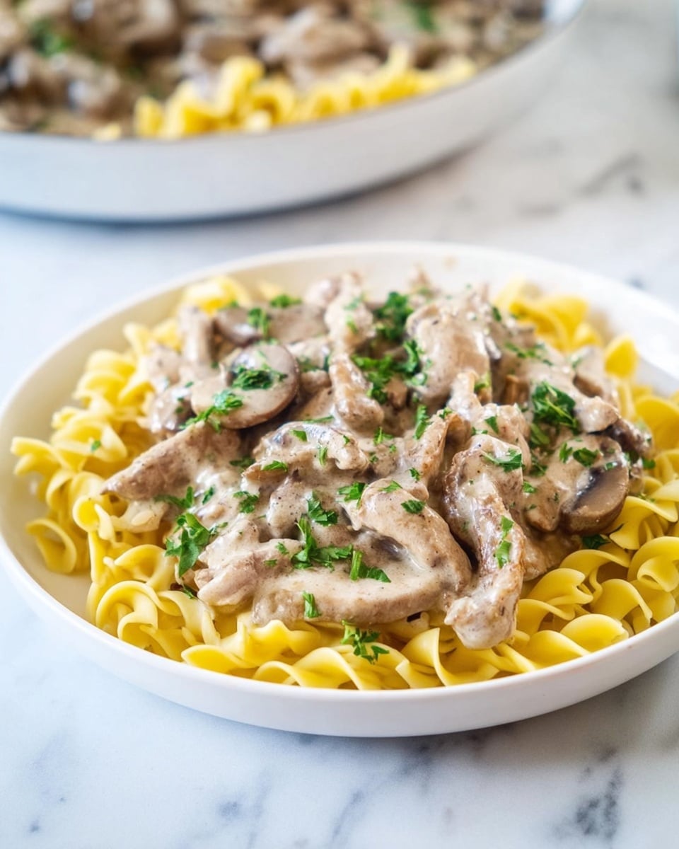 A white plate sits on a white marbled surface, filled with three layers of food. The bottom layer is yellow egg noodles, soft and slightly curled. The middle layer has light brown strips of cooked meat mixed with slices of brown mushrooms. The top layer is a creamy, beige sauce that coats the meat and mushrooms, garnished with small green pieces of chopped parsley scattered across the dish. In the background, there is a blurred serving dish with more of the same food. Photo taken with an iphone --ar 4:5 --v 7