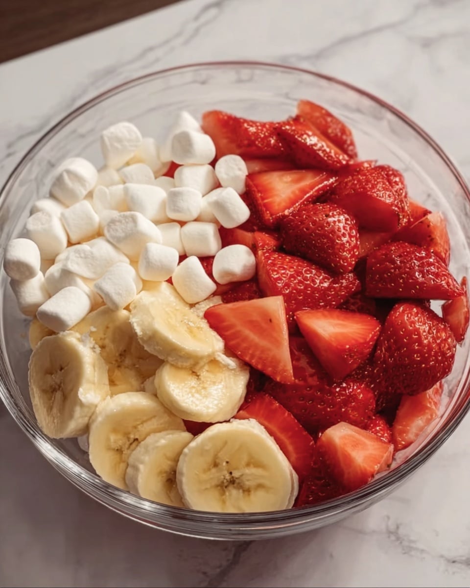 A clear glass bowl is filled with three main layers side by side: one section is small white marshmallows, next to it is a pile of sliced pale yellow bananas, and the largest portion is bright red sliced strawberries with a juicy texture. The bowl sits on a white marbled surface. Photo taken with an iphone --ar 4:5 --v 7