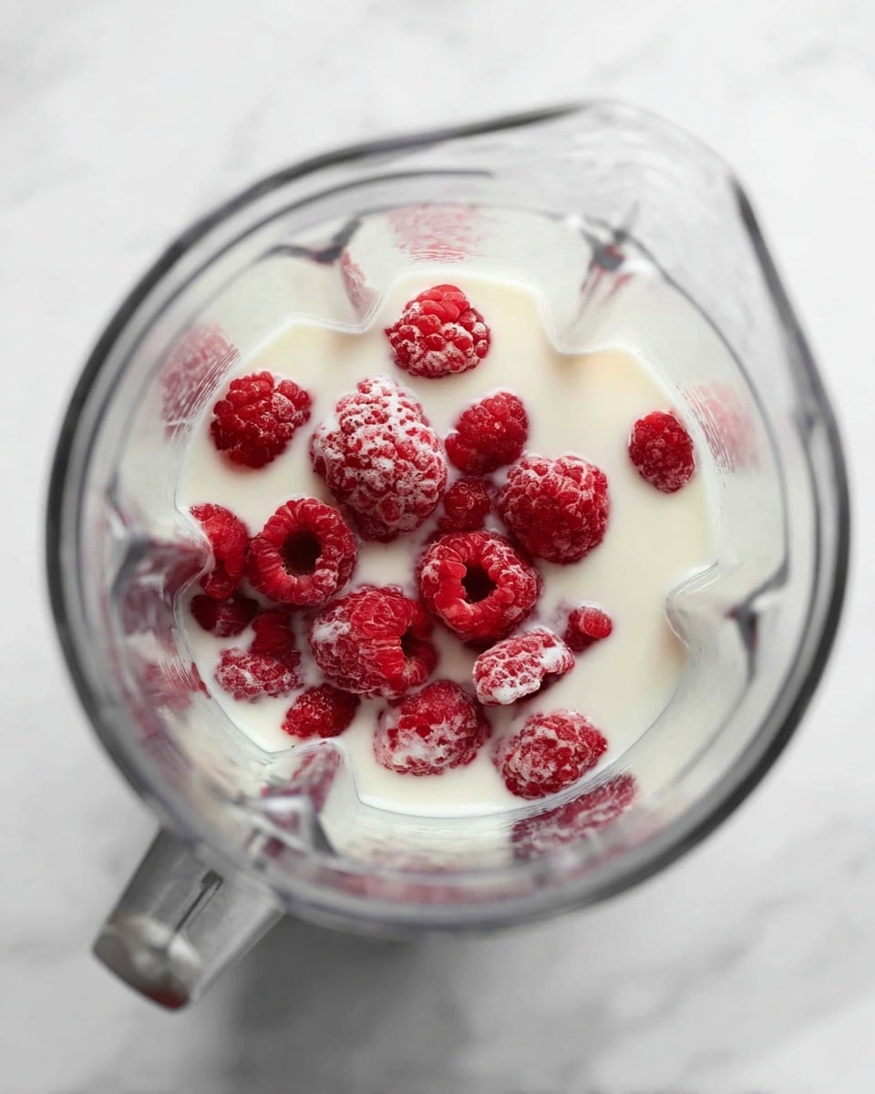 A clear blender jar with a mix of layers inside: the bottom layer is white milk with a smooth texture, floating on the surface are several red raspberries, some fully submerged and others partially, showing their bumpy texture. The blender sits on a white marbled surface, and the image is taken from above, showing the clear sides of the blender and the colorful raspberries against the white milk in a soft natural light. photo taken with an iphone --ar 4:5 --v 7
