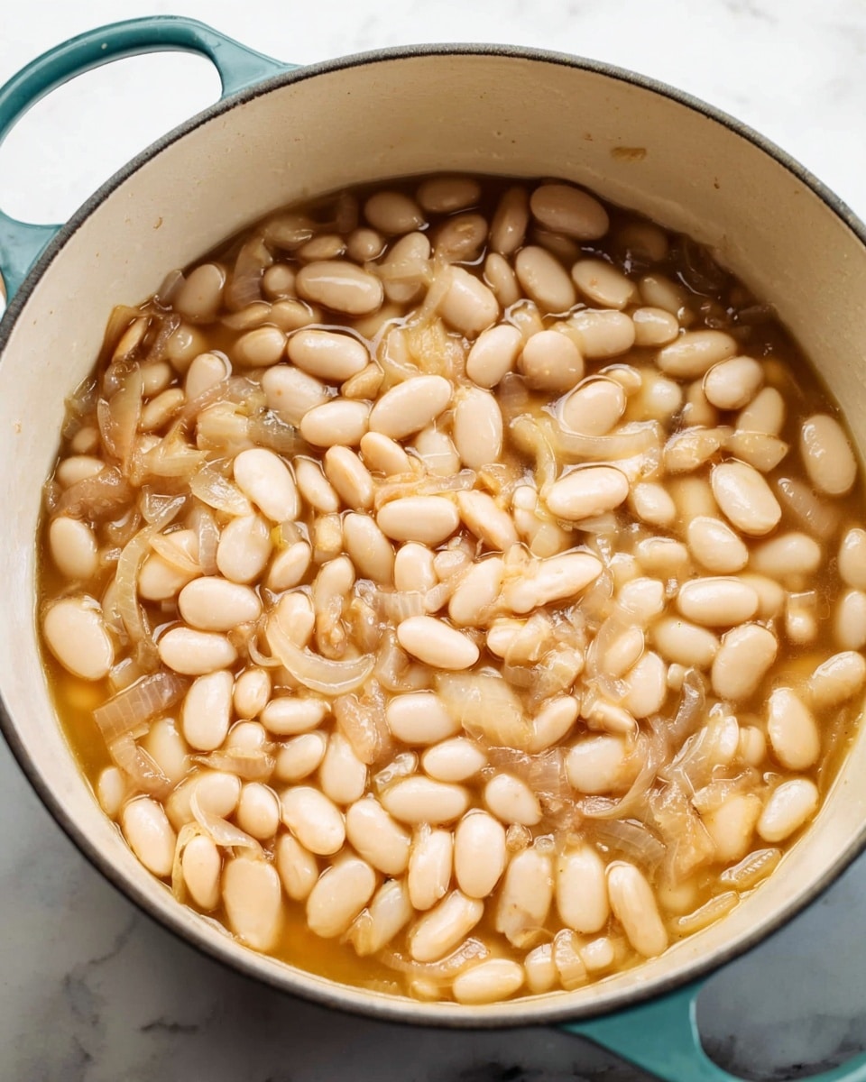 A close-up of a large white pot with teal handles filled with cooked white beans in light brown broth, with thin slices of translucent shallots mixed throughout. The beans are plump and soft-looking, layered evenly across the pot in a shallow pool of broth with slight shiny reflections. The pot sits on a white marbled surface, giving a clean and bright kitchen feel. Photo taken with an iphone --ar 4:5 --v 7