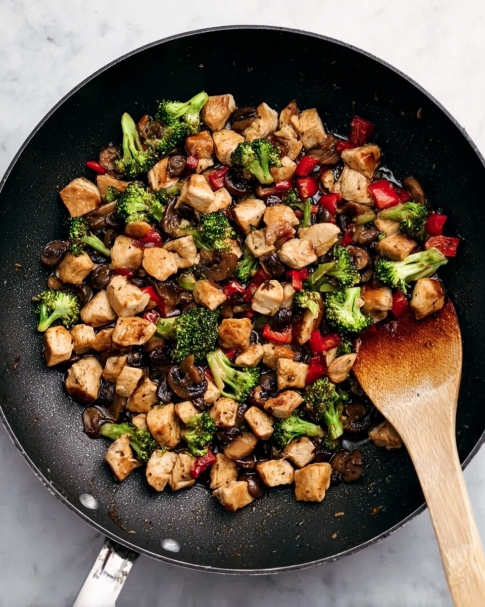 A black pan filled with small pieces of cooked chicken, bright green broccoli florets, dark brown mushrooms, and small red bell pepper cubes, all mixed together and slightly shiny with oil. A wooden spatula rests on the right side of the pan which sits on a white marbled surface. The scene shows a simple colorful stir-fry with bits of each ingredient evenly spread across the pan. photo taken with an iphone --ar 4:5 --v 7