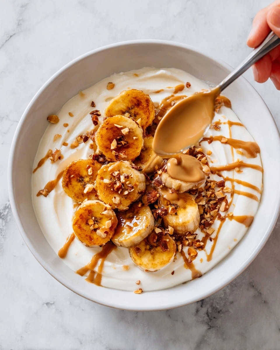 A white bowl with a base layer of smooth, creamy white yogurt spread around the bowl. On top are two layers of golden brown caramelized banana slices arranged in a slightly random cluster in the center. Drizzled over the bananas is a light brown caramel or peanut sauce in thin streams. Scattered around the bananas are small pieces of crushed nuts adding a rough texture contrast. A spoon held by a woman’s hand is pouring more of the light brown sauce over the bananas. The bowl sits on a white marbled surface. photo taken with an iphone --ar 4:5 --v 7