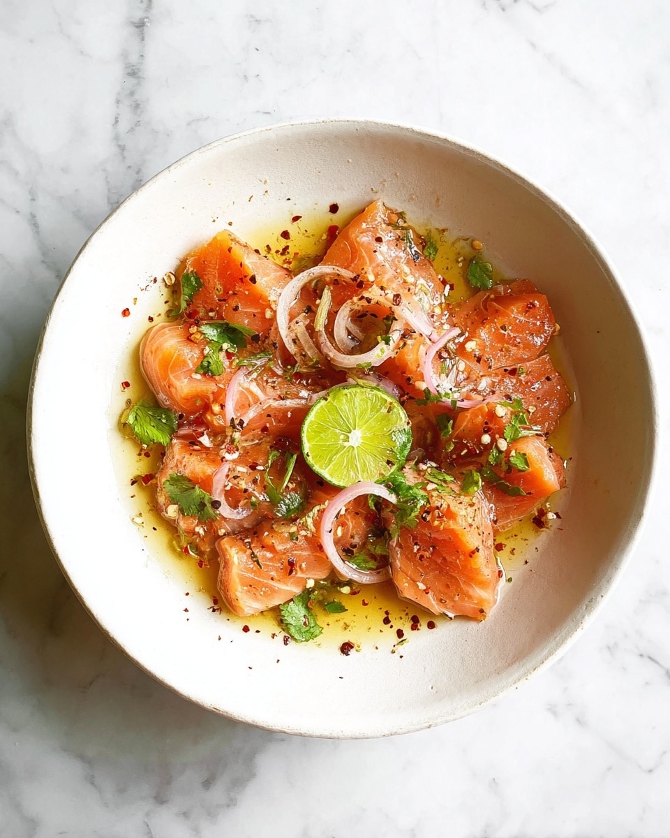 A white bowl on a white marbled surface holds thin, folded slices of orange-pink salmon, arranged loosely in a single layer with some overlapping. Scattered on top of the salmon are thin, pale rings of shallots, small green cilantro leaves, and a drizzle of golden olive oil with specks of black pepper and red chili flakes that add texture and color contrast. The salmon looks fresh and slightly shiny from the oil, with a lightly marbled texture visible on the fish. photo taken with an iphone --ar 4:5 --v 7