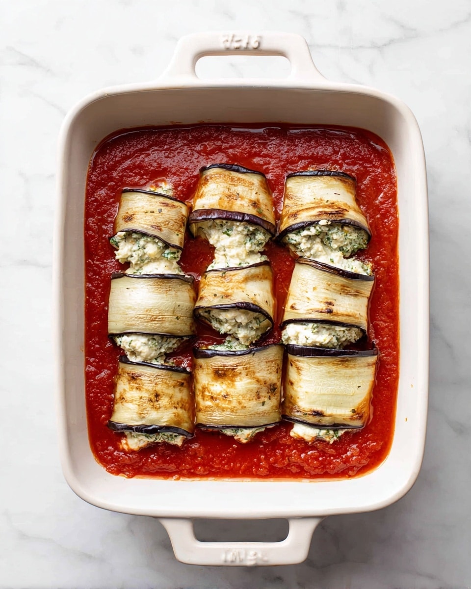 A white square baking dish with handles holds six rolled eggplant slices filled with a creamy cheese and herb mixture, arranged in two rows on a smooth layer of thick, bright red tomato sauce. The eggplant slices show light brown grill marks and slight purple edges, with the filling peeking out at the ends. The dish sits on a white marbled surface. photo taken with an iphone --ar 4:5 --v 7