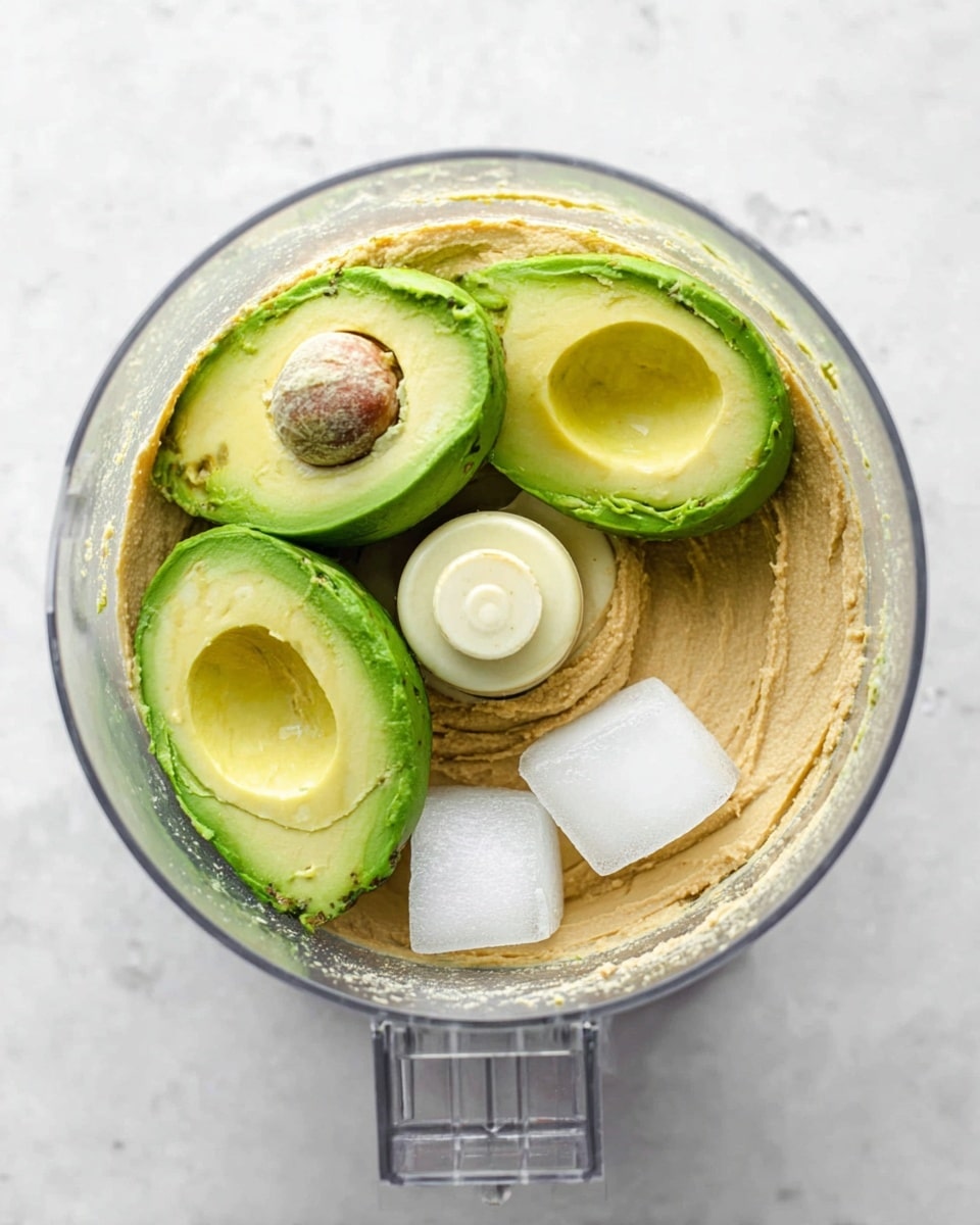 A clear food processor bowl holds a creamy beige base layer of smooth hummus spread at the bottom, with three large green avocado halves placed on top showing different textures of smooth and slightly rough surfaces. Nestled near the avocados are three white ice cubes that add a sharp contrast in texture and color. The clear plastic bowl sits on a white marbled surface, and the photo is taken from above to capture all the details. photo taken with an iphone --ar 4:5 --v 7